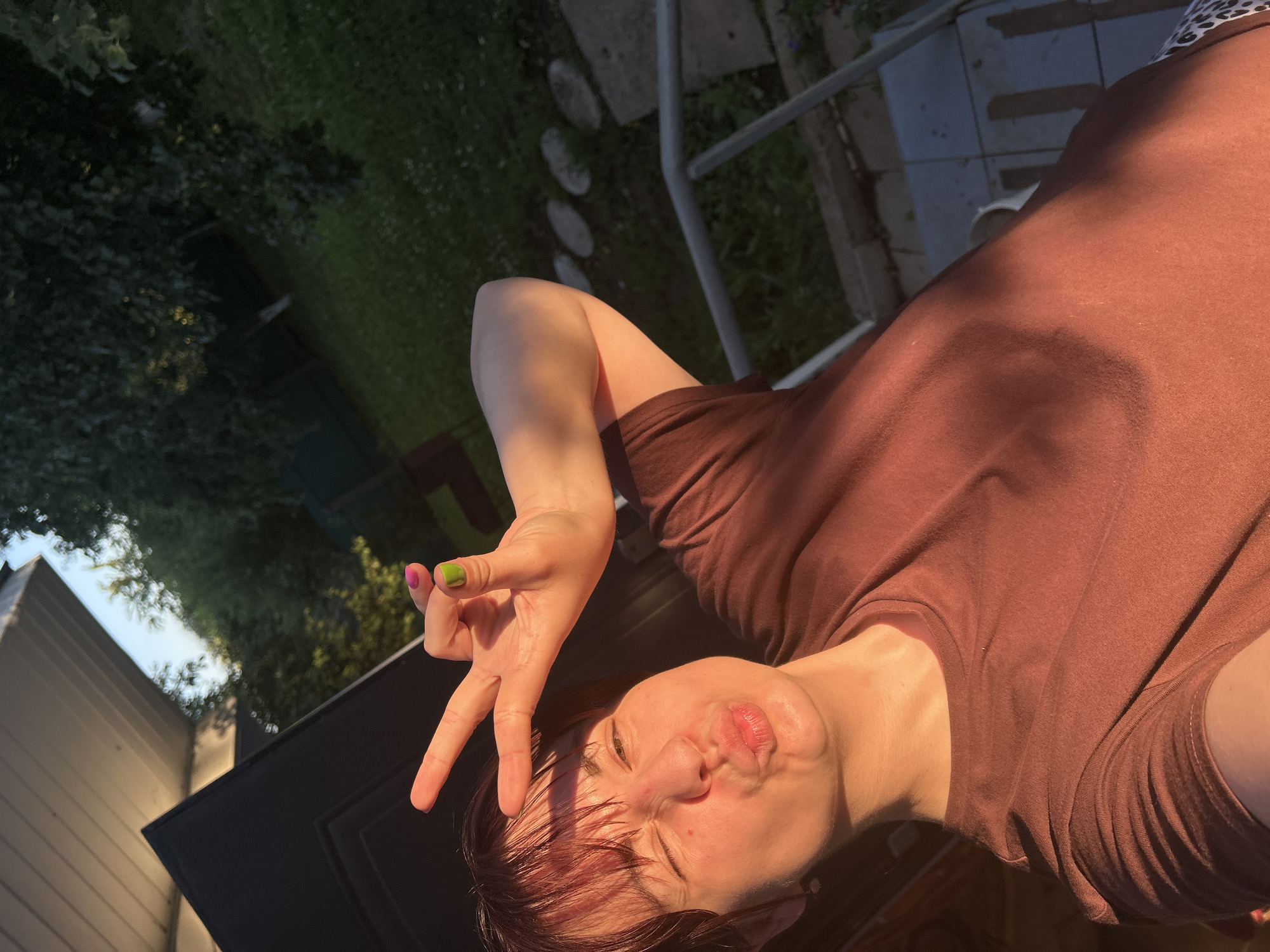 A young woman with dark hair and bangs is taking a selfie outdoors in what appears to be a backyard. She is wearing a brown t-shirt and has her right hand raised, making a peace sign with her fingers. Her lips are pursed, and she is winking. The sun is setting, casting a warm glow on her face and arm. In the background, there is green grass, trees, and a wooden structure. The image is taken in the city of Fokino, Russia, as indicated by the prompt.