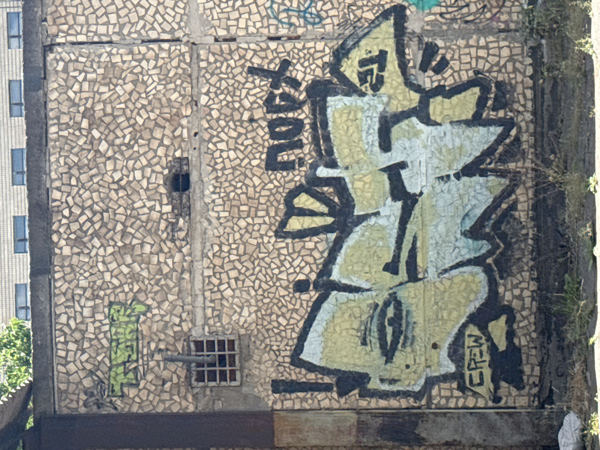 An outdoor, medium shot shows a wall covered in small, tan, irregularly shaped tiles, with various graffiti tags visible. The graffiti includes a green tag on the upper left with "TSHR" written in block letters. Below this, a large, multi-colored graffiti piece spans most of the wall, featuring yellow, light blue, and black elements. Inside this piece, "CREW" is written in black block letters on the lower left. Above the main graffiti, to the right, is a black tag that reads "nocx". The wall also has a small, barred window and a pipe. The ground in front of the wall is covered in dry grass and debris. The background shows a building with windows and a green tree.