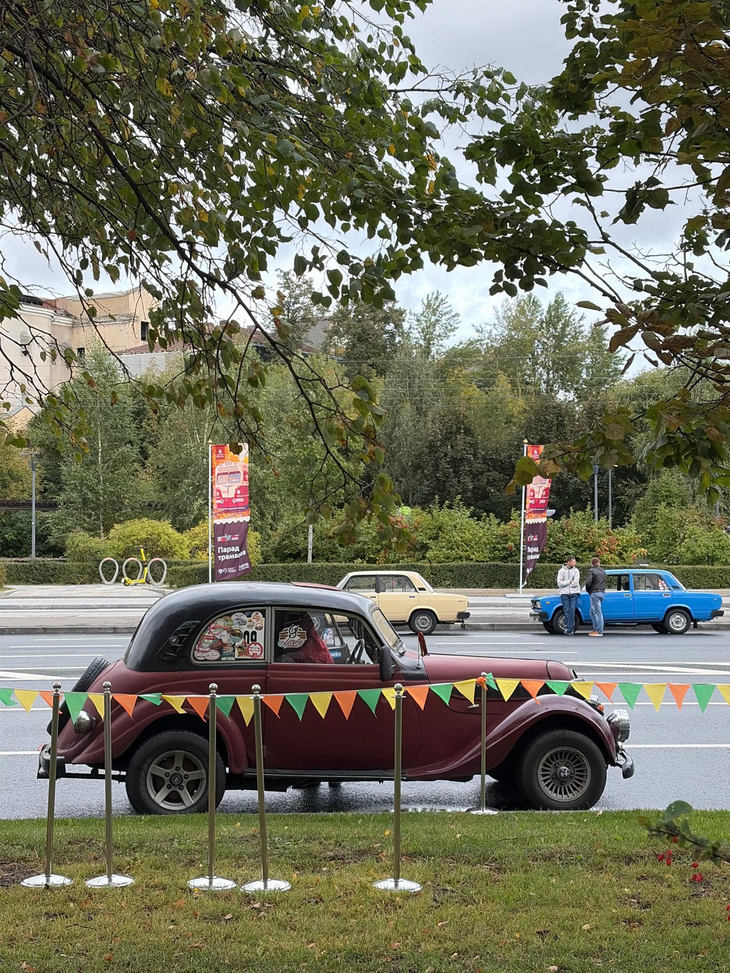 This image captures a street scene in Moscow, Russia, with several vintage cars parked along the roadside. In the foreground, a burgundy-colored vintage car is prominently displayed, adorned with various stickers on its rear window. A decorative string of colorful triangular flags is strung across stanchions in front of the car, creating a visual barrier.
Further down the street, a pale yellow vintage car is visible, followed by a bright blue sedan. Two men are standing near the blue car, engaged in conversation.
In the background, there are trees and a building, suggesting an urban environment. Two tall banners, likely advertisements or event signage, are placed strategically along the street. One banner features an image of a retro tram and text in Cyrillic, possibly indicating a transportation-themed event or exhibition. A unique sculpture resembling three interconnected bicycle wheels is also present on the grass verge, adding an artistic element to the scene. The sky is overcast, indicating a cloudy day.