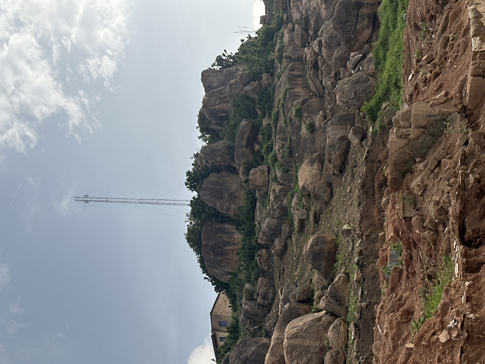 The image depicts a rocky, overgrown hillside with a telecommunications tower and a building visible on top. The terrain is uneven, characterized by large boulders, sparse vegetation, and reddish-brown soil. A structure with a light-colored facade and a few windows is situated on the upper part of the hill, to the left of the telecommunications tower. The tower itself is a tall, lattice-style structure. The sky above is partly cloudy, with a bright, diffused light suggesting daytime. The foreground shows a steep, eroded bank with more rocks and scattered debris. The scene appears to be an outdoor environment, possibly in a semi-urban or rural setting, given the presence of the tower and building amidst the natural landscape.