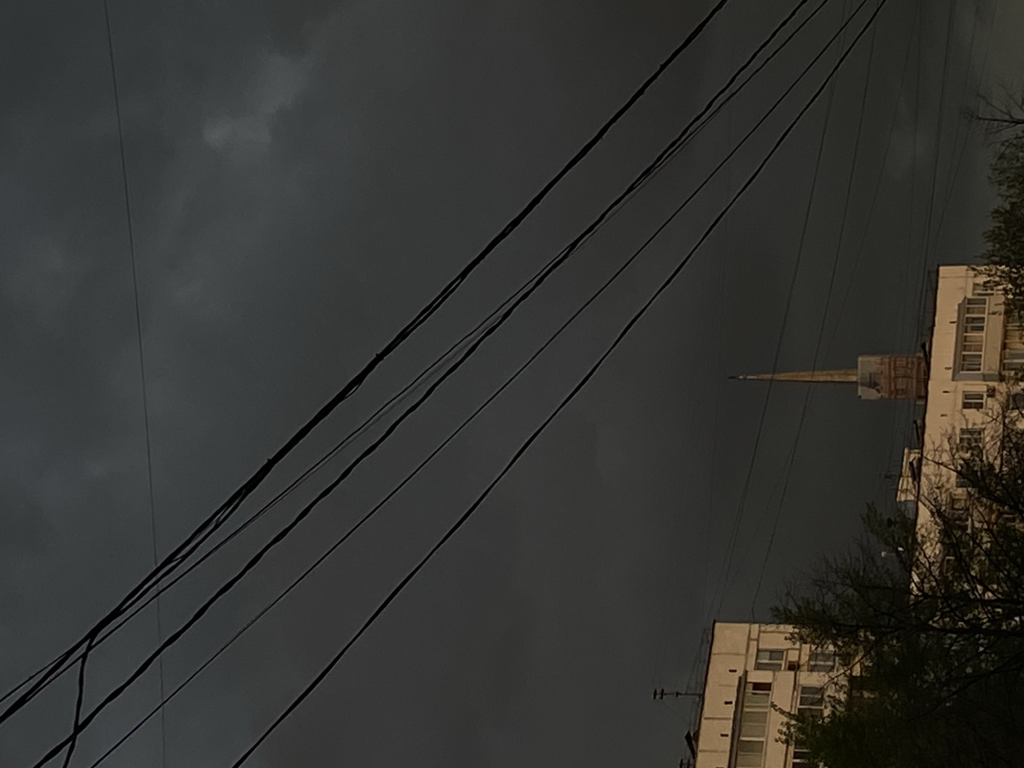 The image depicts an urban or suburban scene under a heavy, dark grey sky, indicative of gloomy weather or an approaching storm. Several thick, black utility lines or power cables stretch diagonally from the upper left portion of the frame towards the lower right.
On the right side of the image, the facades of two multi-story residential buildings are visible. The buildings are made of light-colored materials, with windows discernable on the more prominent, lower building. Behind these structures, a tall, slender, tapering spire or tower with a pointed top rises sharply against the dark sky, suggesting a significant landmark or architectural feature, possibly in Moscow, Russia as per the context.
Bare tree branches, mostly devoid of leaves, are present in the bottom right corner, partially obscuring the lower building. No human figures or discernible activity are captured in the frame. The time of day appears to be either late afternoon/early evening or a deeply overcast day, contributing to a somber and tranquil atmosphere. No text is visible in the image.