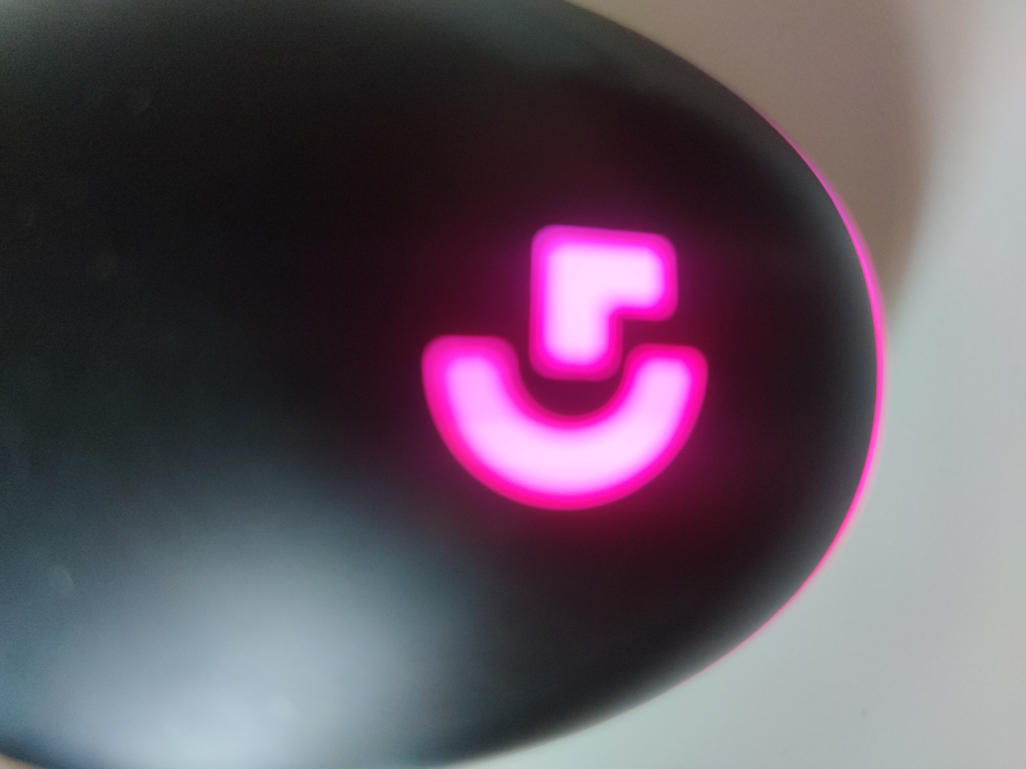 A close-up, out-of-focus image shows the logo of a Logitech device, possibly a mouse. The logo is illuminated in a bright pink color and set against a dark, possibly black or grey, surface. A portion of the device's curved edge is visible, with a slightly brighter area in the background suggesting a light source. The location is unknown.