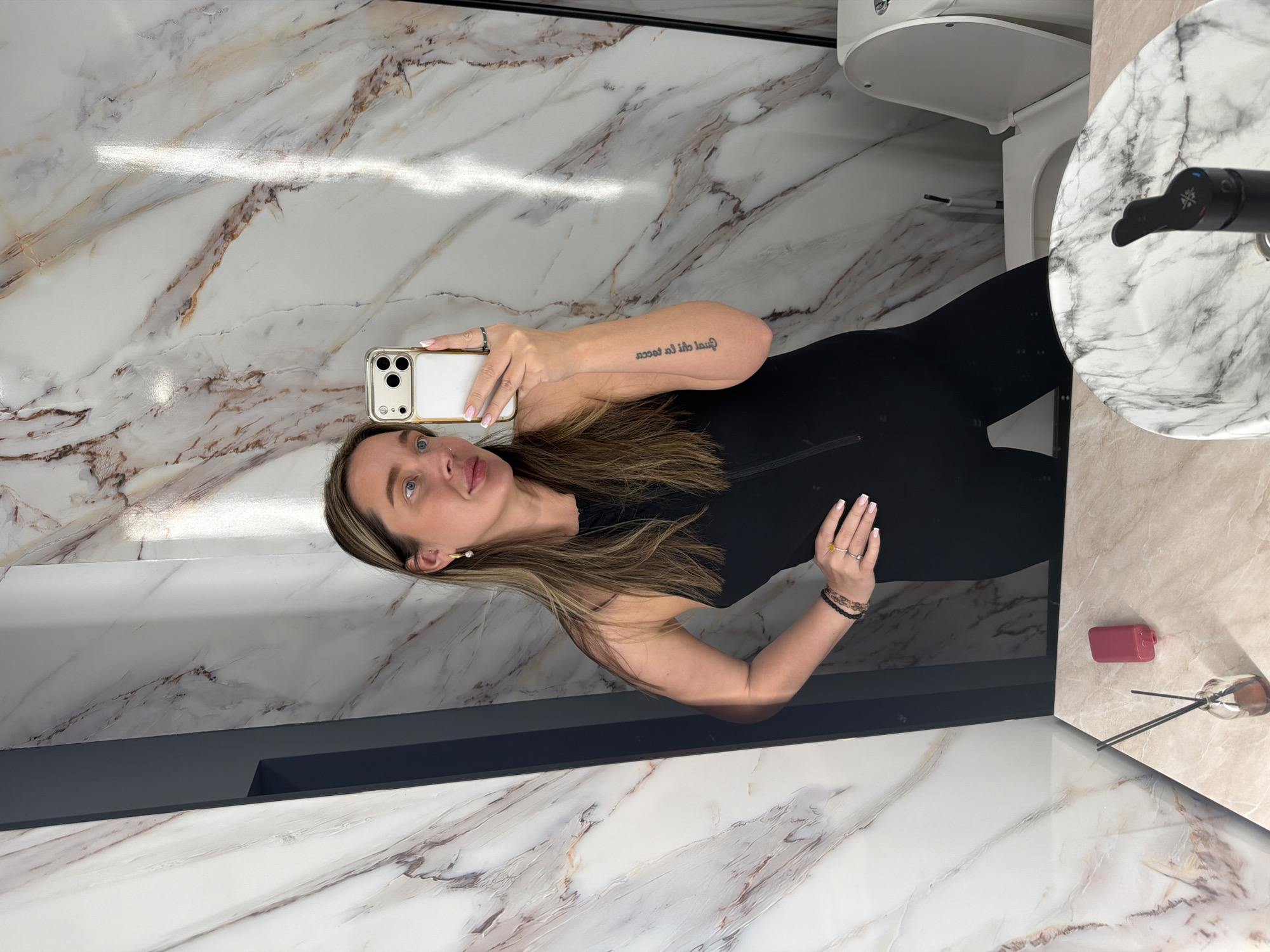The image shows a woman taking a selfie in what appears to be a bathroom. The woman is holding a smartphone with a white and gold case. She is wearing a black top. Her hair is light brown with blonde highlights. She has a tattoo on her upper left arm. The bathroom has white marble walls and countertops. There is a white toilet and sink visible. A diffuser and a pink container are on the counter. There is a dark grey trim accenting the space. There is nothing in the image to indicate the location is Moscow, Russia.