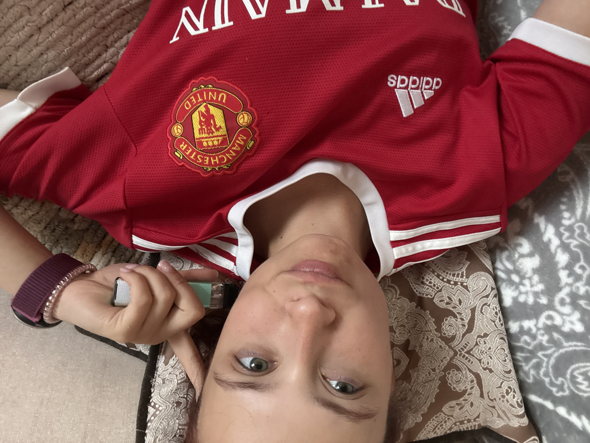 A person lies on a patterned pillow, wearing a red Manchester United soccer jersey with Adidas logo, holding a lighter in their hand. The person is positioned within the city of Прикамский, Russia. They wear a burgundy Apple Watch band and a beaded bracelet.