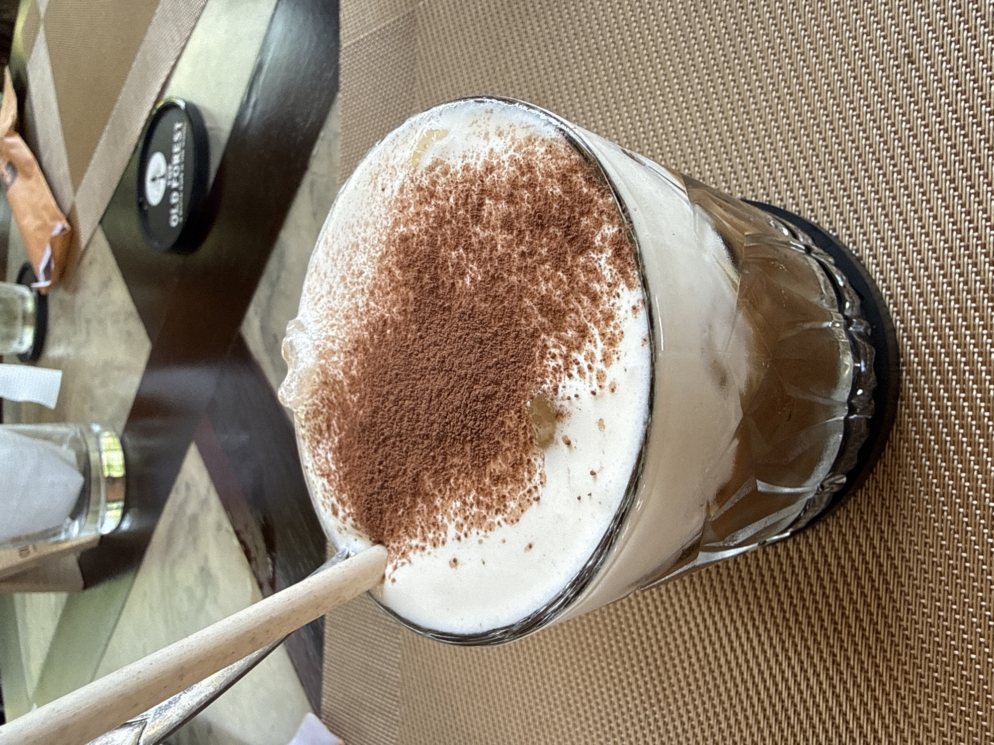 The image displays a close-up, high-angle view of an iced beverage on a table, captured in Phú Quốc, Vietnam. The central object is a tall glass of a brown-colored drink, likely iced coffee, generously topped with a thick layer of white foam and a sprinkling of dark brown cocoa powder. A light-colored straw, appearing to be made of wood or paper, is inserted into the drink.
The beverage rests on a light brown woven placemat, which covers a table featuring a dark and light brown geometric pattern. To the left of the glass, a small, black cylindrical container is visible, bearing the white text "OLD FOREST" and additional, smaller text below it. Further to the left, parts of a crinkled brown paper packet and a small glass shaker can be seen. The overall setting suggests a calm, daytime environment, possibly a cafe or restaurant, where a refreshing drink is prepared for consumption. No people are visible, and the activity implied is the enjoyment of the beverage.