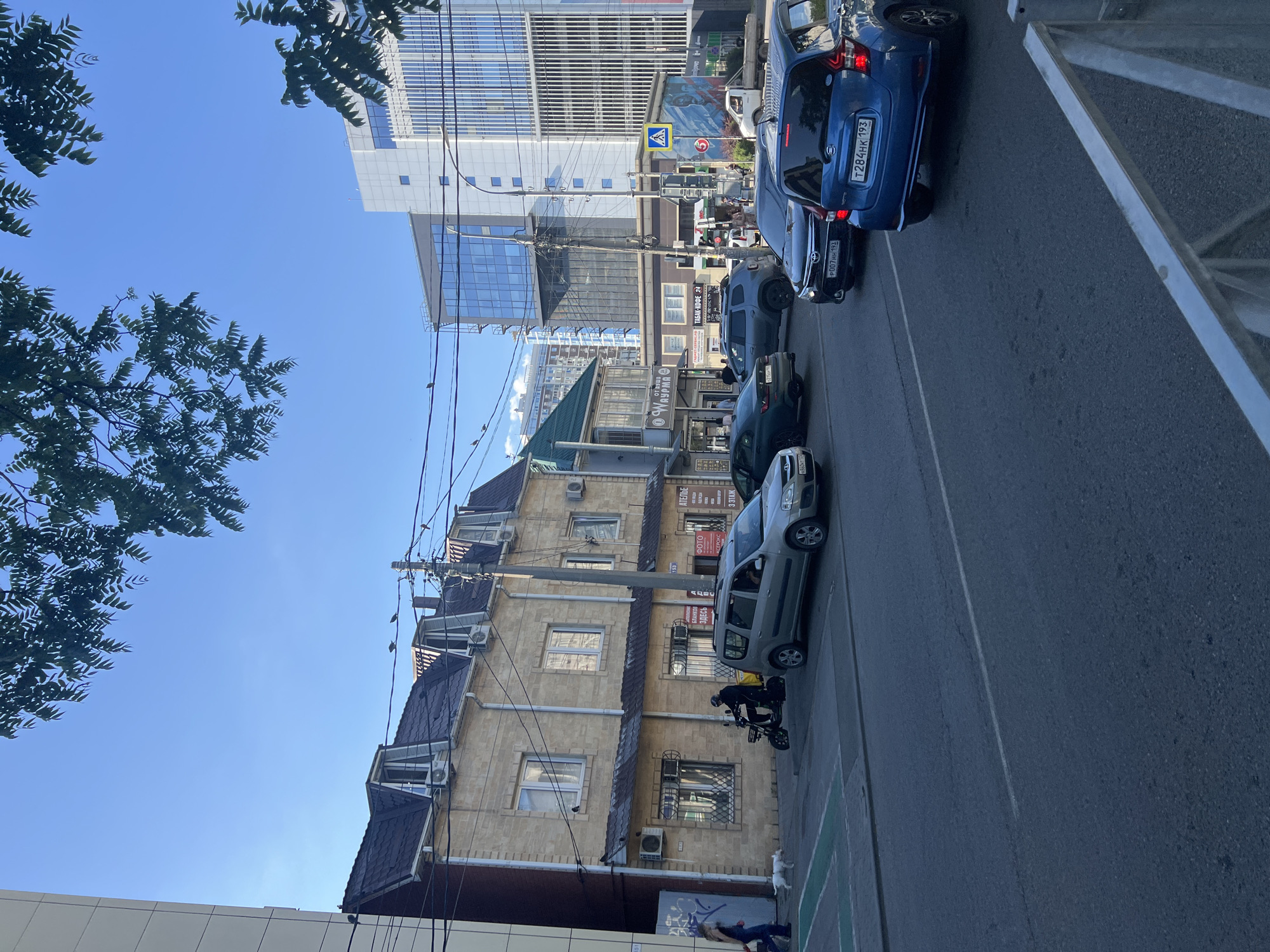 In an urban street scene, a two-story building with a light-colored brick facade and a dark-tiled roof stands on the left, featuring several dormer windows and air conditioning units. A pedestrian, a person in dark clothing, walks with a small white dog on the sidewalk in front of this building. To the right of the pedestrian, a person wearing a helmet rides a scooter. Adjacent to the scooter, a light-colored minivan is parked.
Further to the right, the street is populated with several cars. A dark-colored SUV is visible, followed by a dark sedan, and then a blue hatchback in the foreground. In the background, modern multi-story buildings with glass facades dominate the skyline, contrasting with the older architecture. A traffic light with a pedestrian crossing symbol is present on the right side of the intersection. Trees with green foliage hang over the scene from the top. The sky is clear and blue.