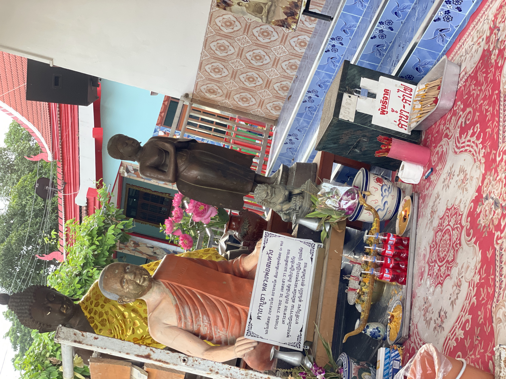 In Pattaya City, Thailand, an outdoor shrine is depicted. The shrine features multiple Buddha statues, some of which are partially draped in orange and yellow fabric. A sign with Thai script is placed in front of a seated monk statue. Various offerings are laid out on a table, including trays of food, several red bottles of Fanta, and incense sticks. Behind the shrine, blue-tiled stairs ascend, and a red-tiled roof of a building is visible. The scene is set against a backdrop of green foliage and a pale blue wall, suggesting an outdoor temple or shrine setting.