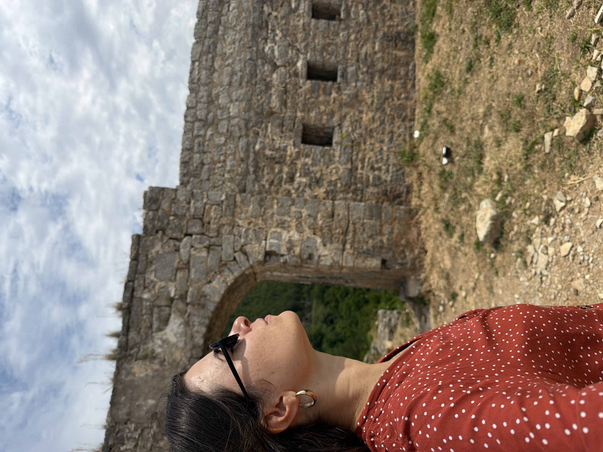 The image is oriented on its side, depicting a person in profile looking upwards towards a cloudy sky. The subject, a woman, is in the lower right, with her face angled towards the left. She has dark hair, wears dark sunglasses pushed onto her head, and a distinctive gold-colored hoop earring. Her top is a red-orange fabric with small white polka dots.
In the background, a substantial ancient stone wall, part of a ruin or fort, occupies the left and central portions of the frame. A large arched gateway is visible in the lower center of the wall, revealing lush green trees and foliage beyond. Above the arch and to the right, there are three small, rectangular openings, stacked vertically within the stone structure. The masonry is rough and uneven, consistent with historic construction.
The ground around the ruin, visible on the right, is dry and earthen, scattered with sparse patches of grass and small rocks. The sky is bright with white, fluffy clouds, indicating daytime conditions. There is no visible text in the image. The scene suggests a visit to a historical site in Budva, Montenegro.