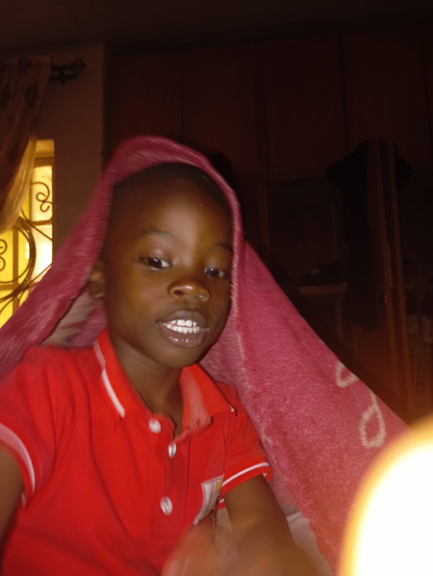 The image shows a close-up, head-and-shoulders shot of a young Black child wearing a red collared shirt with white trim. The child is looking directly at the camera with an open mouth, revealing white teeth. A pink blanket or towel with a white circular pattern is draped over the child's head and shoulders, partially obscuring the background. The background appears to be an indoor domestic setting, possibly a bedroom, with dark wooden cabinets or shelves visible. To the left, a window with a decorative metal grate and a yellow light source is partially visible. The lighting in the image is dim, with a bright, circular light source in the lower right corner, likely a camera flash. There are no discernible details indicating a specific location within Jos, Nigeria, beyond it being an indoor residential environment.