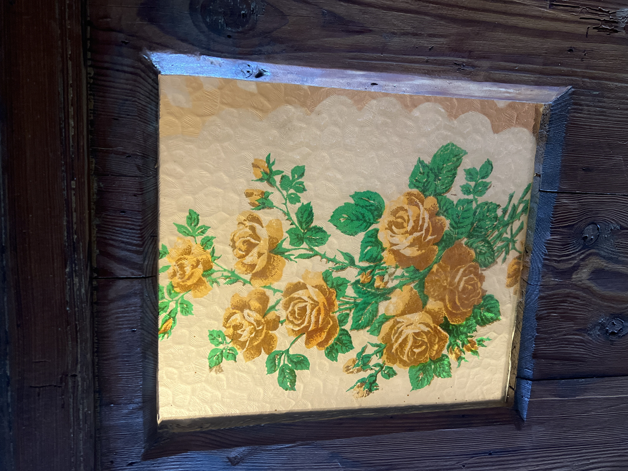 The image shows a close-up of a decorative panel with a floral pattern, specifically yellow roses and green leaves, set within a wooden frame. The panel appears to be made of textured glass or a similar material, with a slightly opaque, patterned background behind the artwork. The wooden frame is dark and appears aged, with visible grain and some imperfections. The overall style suggests it might be a decorative element on a piece of furniture or a door. The context provided places this object in Norviliškės, Lithuania, suggesting it's an indoor decorative detail within a building in that location. There are no people, actions, or interactions depicted in the image.