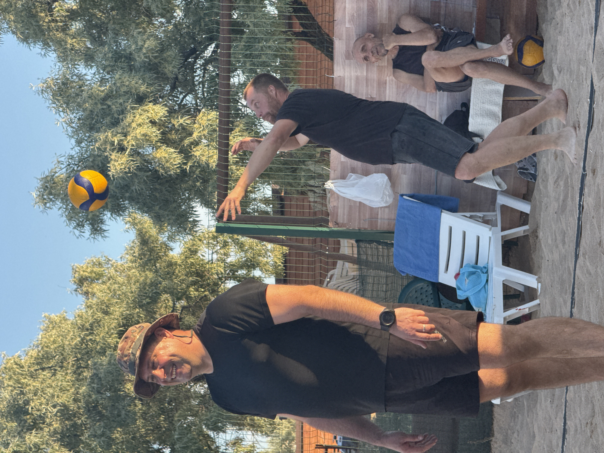 Three adult males are on a sandy beach, seemingly playing or watching a volleyball game. The man in the foreground on the left is wearing a camouflage hat, a black t-shirt, and dark shorts. He is smiling and looking to his left. A smartwatch is visible on his left wrist. In the middle of the frame, a man in a black t-shirt and dark shorts is in mid-action, reaching up towards a volleyball in the air. He is looking up at the ball. To the right, another man, appearing older, is seated on a wooden bench, wearing a dark tank top and shorts. He is looking towards the volleyball game, with his chin resting on his hand. A volleyball, yellow and blue, is suspended in the air between the two active men. Behind them, a fence, trees, and lounge chairs are visible. The setting suggests an outdoor recreational area.
