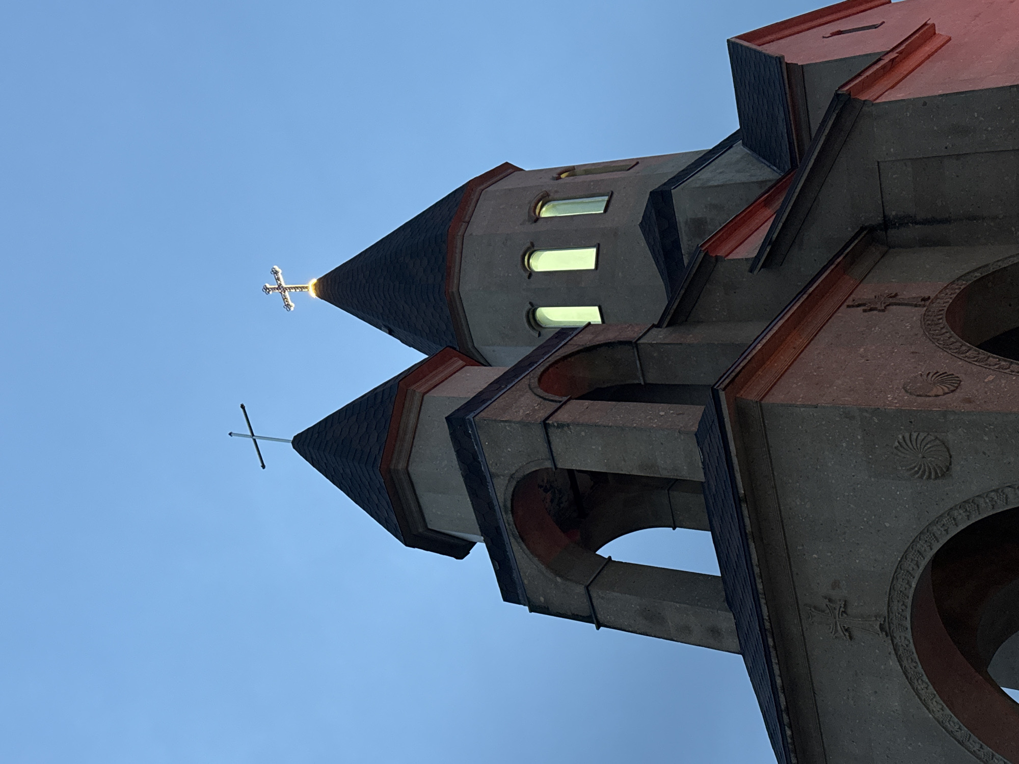 The image shows a low-angle view of a church structure against a clear blue sky. The church is constructed of gray stone with reddish-brown accents. It features two prominent towers with conical roofs, each topped with a cross. The tower on the left has a simple cross, while the tower on the right has a more ornate, glowing cross. The structure includes architectural details such as arched openings, decorative crosses, and geometric patterns on the stone surface. The scene is likely in Novomikhaylovsky, Russia, given the prompt's reference to that location.