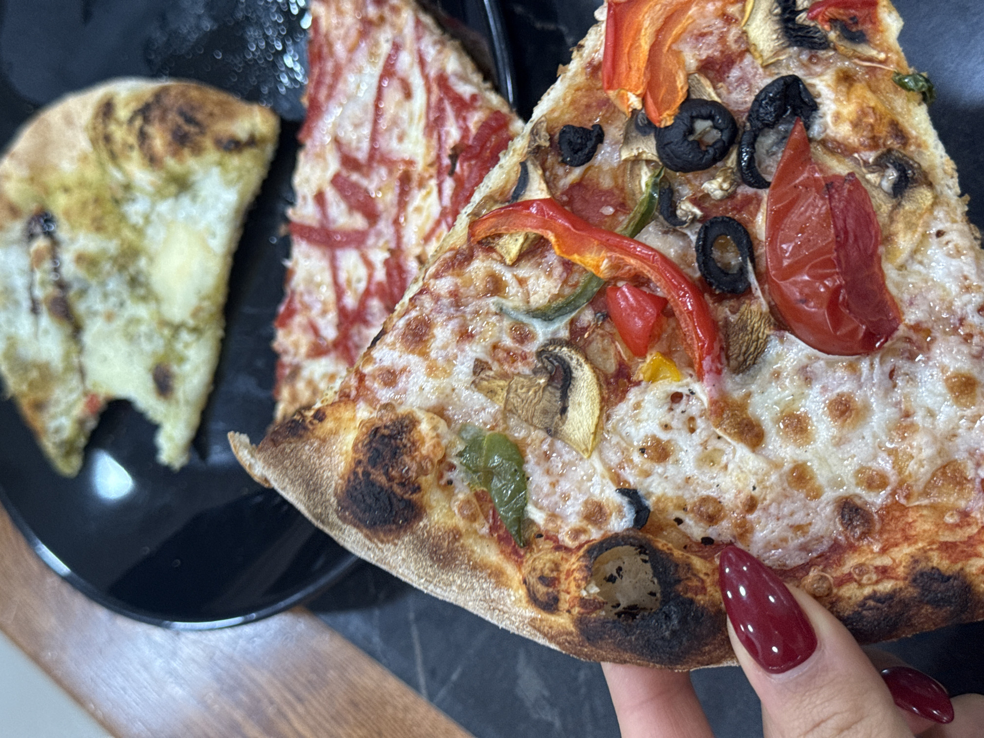 A close-up photograph shows three slices of pizza on a black plate. One slice has green pesto, the second has red sauce in lines, and the third is topped with mozzarella cheese, red and yellow bell peppers, sliced mushrooms, black olives, and a tomato. The photograph features a hand with red polished nails holding the slice with multiple toppings. The background appears to be a wooden surface. The photograph does not provide any information to definitively conclude that the photo was taken in the city of Aparan, Armenia.