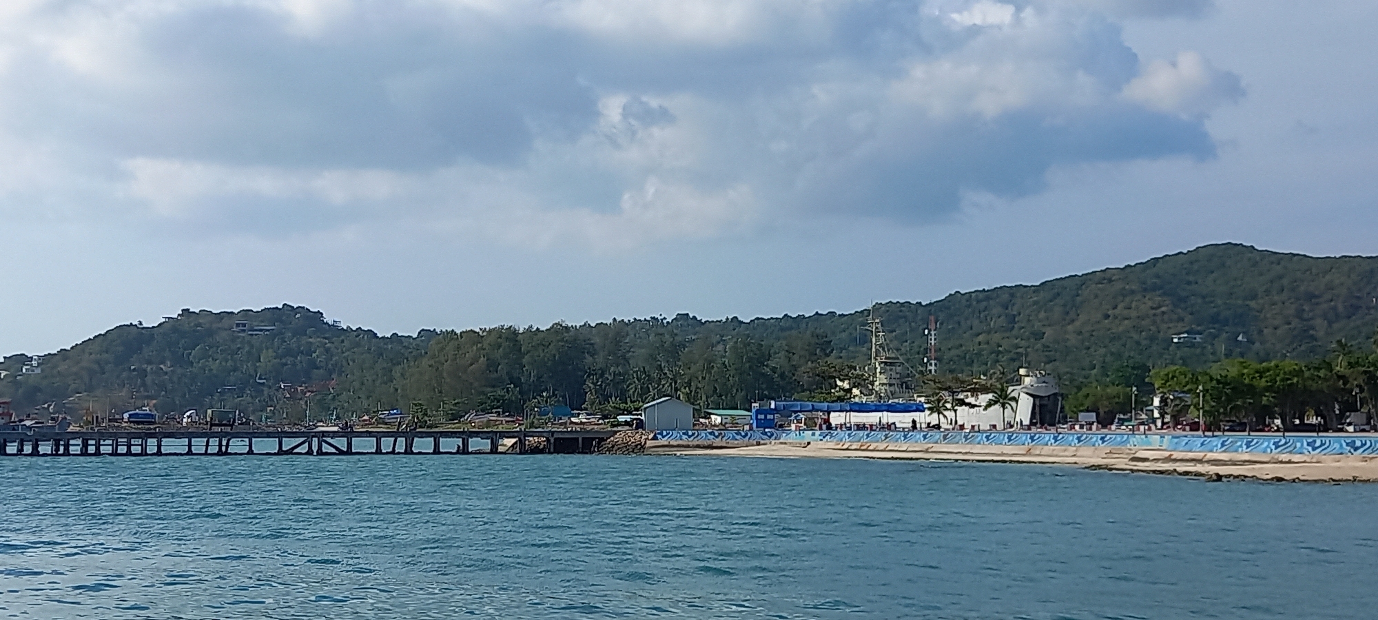 The image presents a panoramic view of a coastal scene in Thong Sala, Thailand, under an overcast sky. The foreground is dominated by the blue-green waters of the sea, showing gentle ripples on its surface.
A long, grey pier, constructed from concrete or wood, extends from the left side of the frame into the mid-ground. Along and behind the pier, several small, indistinct boats or fishing vessels are visible. Beyond the pier and continuing to the right, a shoreline transitions from a narrow beach to a constructed waterfront.
Along this waterfront, various structures are present. A distinctive blue and white patterned seawall or promenade runs parallel to the shore on the right side, with a road and a few vehicles visible behind it. Several buildings, including a large white building that might serve as a terminal or administrative facility, are situated along this strip. Further back, tropical vegetation, including palm trees, can be seen.
The background is formed by a series of lush green hills or mountains, densely covered in trees. Scattered structures, possibly houses or resorts, are visible on the slopes and peaks of these hills. On the right side, two prominent towers, one appearing to be a communication tower, rise against the forested hills. The sky above is largely cloudy, suggesting a bright but overcast day. The scene depicts a typical coastal town setting with a mix of natural landscape and human development. No discernible text or prominent human activity is visible.