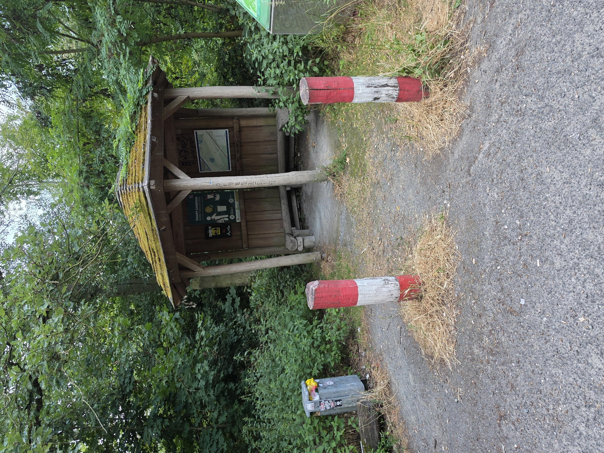 The image depicts an outdoor information kiosk situated within a wooded area. The kiosk is a wooden structure with a moss-covered, multi-pitched roof. It features an open design with wooden posts supporting the roof and has benches inside. Informational signs are mounted on the kiosk walls, including what appears to be a map and text in German. In front of the kiosk, two red and white striped wooden posts stand on a gravel path. To the left of the path, a silver-colored trash can with stickers and a yellow trash bag visible inside is placed near some vegetation. The surrounding environment is dense with green foliage and trees, suggesting a natural or park-like setting. The scene is static, with no people or animals present. The overall impression is that of a rest stop or information point along a trail. This information kiosk is located at the Kelsterbach waterfront.
