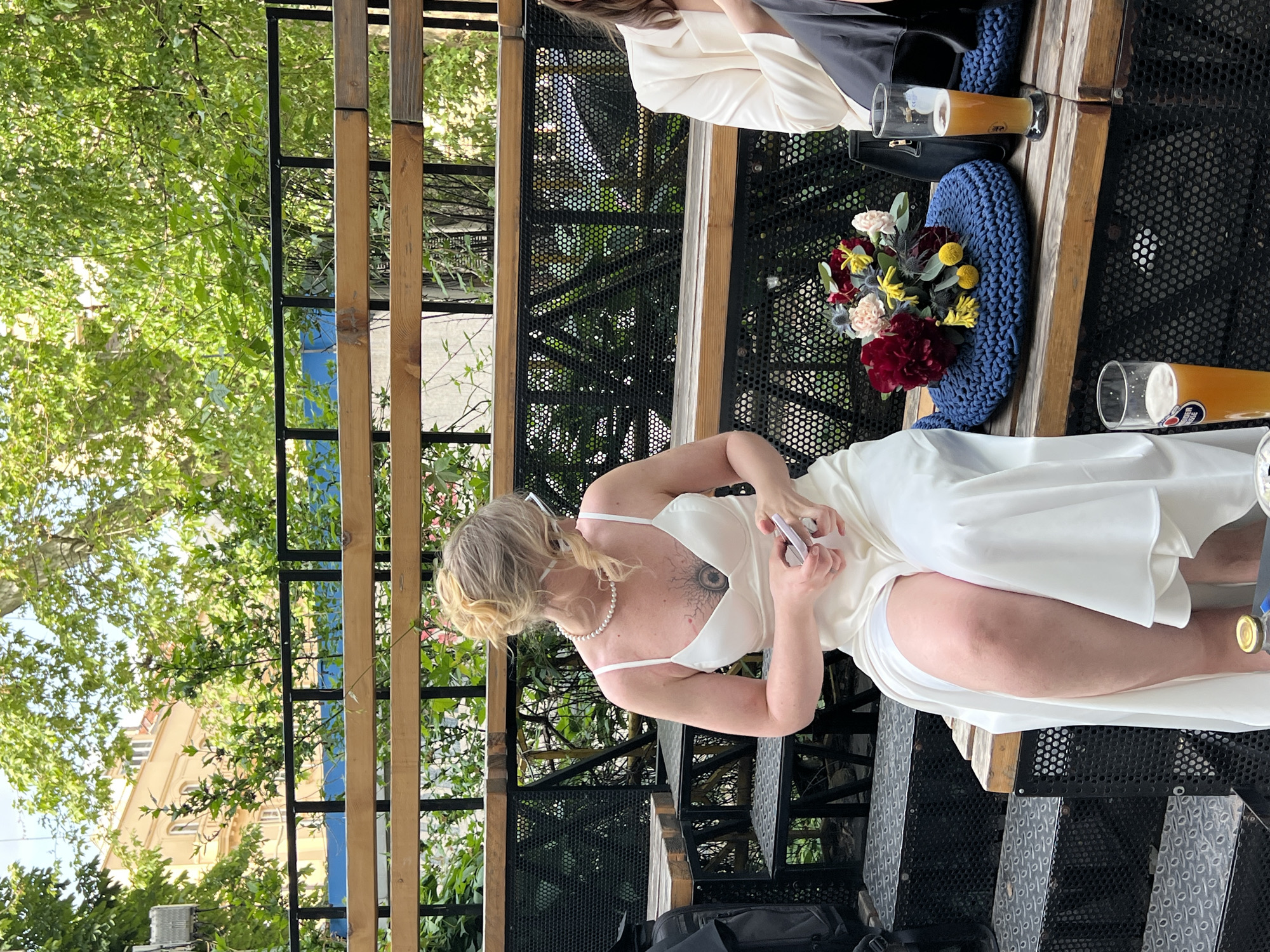 A medium shot, taken from an elevated position, features a woman in a white dress, seated on a bench and holding a mobile phone. The woman has blonde hair and a tattoo on her upper chest/shoulder. She is wearing a pearl necklace.
On the bench next to her is a blue pillow and a floral bouquet. To her right, a beer glass sits on the bench. Another person is seated on the same bench at the upper right.
The location appears to be an outdoor cafe or restaurant. The background consists of trees, buildings, and metal lattice. In the background, a structure can be seen that may be the facade of a building. Due to the style of the trees and architecture, it is possible that this is indeed Belgrade, Serbia.