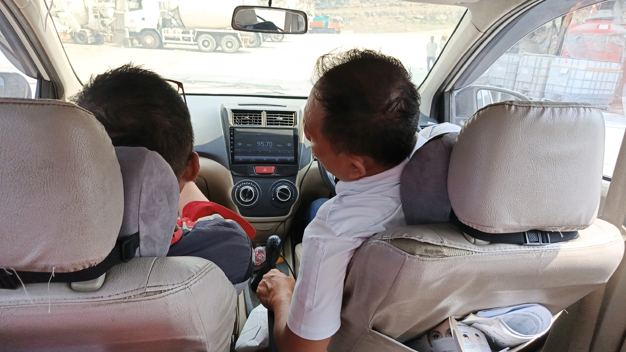 The image is a rear view from inside a car, showing two men and the interior of the vehicle. The man in the front passenger seat is wearing a white collared shirt and is looking towards the dashboard, where a car stereo system is visible displaying "95.70 MHz". The driver, seen from behind, is wearing a dark shirt. The car seats are covered in beige upholstery. Through the windows, a construction site is visible, with concrete mixer trucks and construction materials. The environment suggests an outdoor location, possibly a construction zone in Cijangkar, Indonesia. The man in the driver's seat appears to be operating the car. The overall scene depicts travel within a built environment, likely related to construction or development.