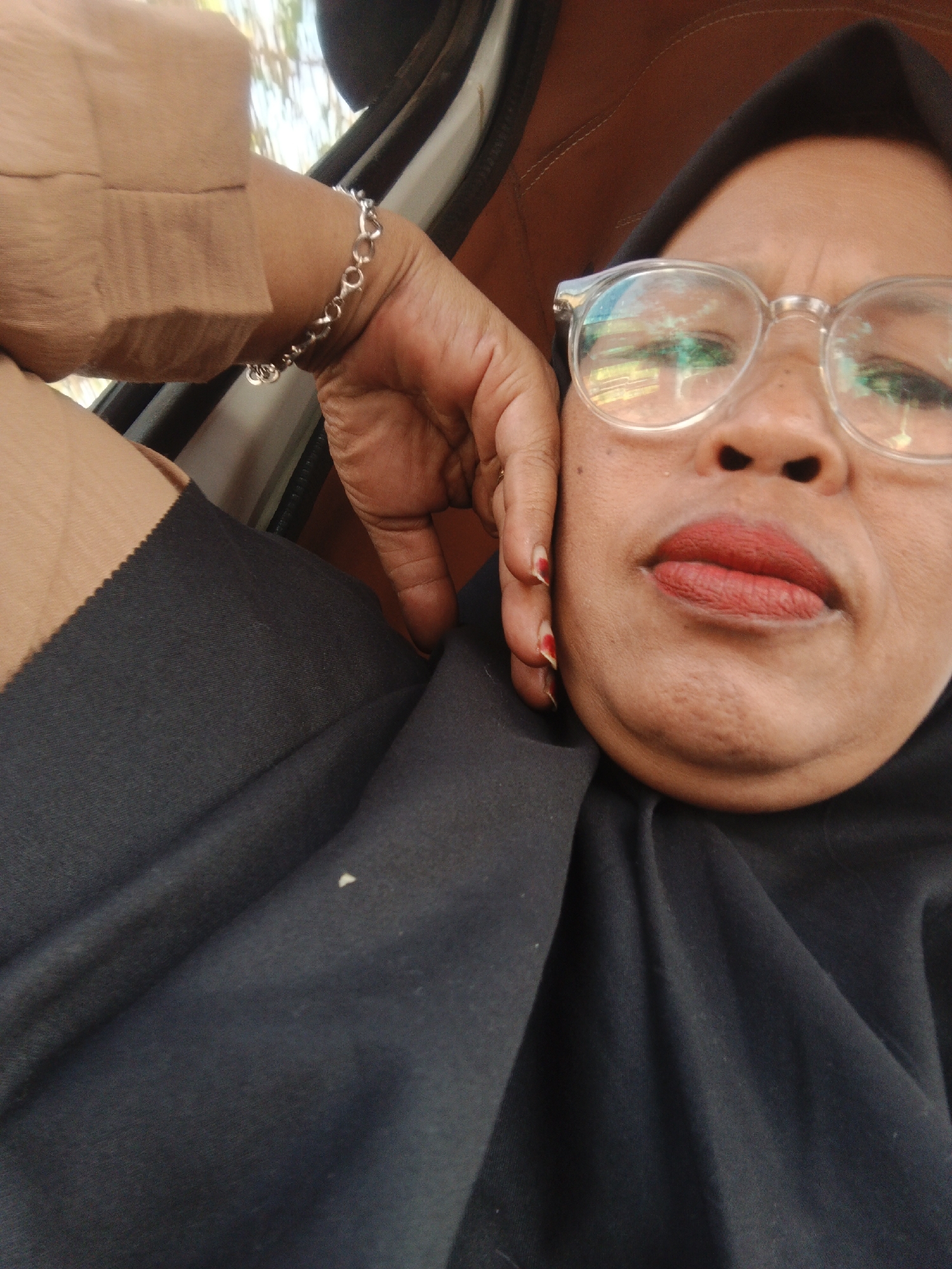 A medium shot captures an individual wearing a black hijab and glasses, indoors. The person is leaning their head on their hand, which rests on their chin, and their arm is draped over their lap, adorned with a silver bracelet. The individual has red lipstick on. The background is blurred, showing what appears to be a car interior with brown upholstery and a window. The image is taken at an angle, with the subject's face filling the frame. The lighting suggests it is daytime, possibly with sunlight reflecting off the glasses, indicating an outdoor environment accessible from the car. The specific location in Sukabumi, Indonesia, cannot be determined from the visual information alone.