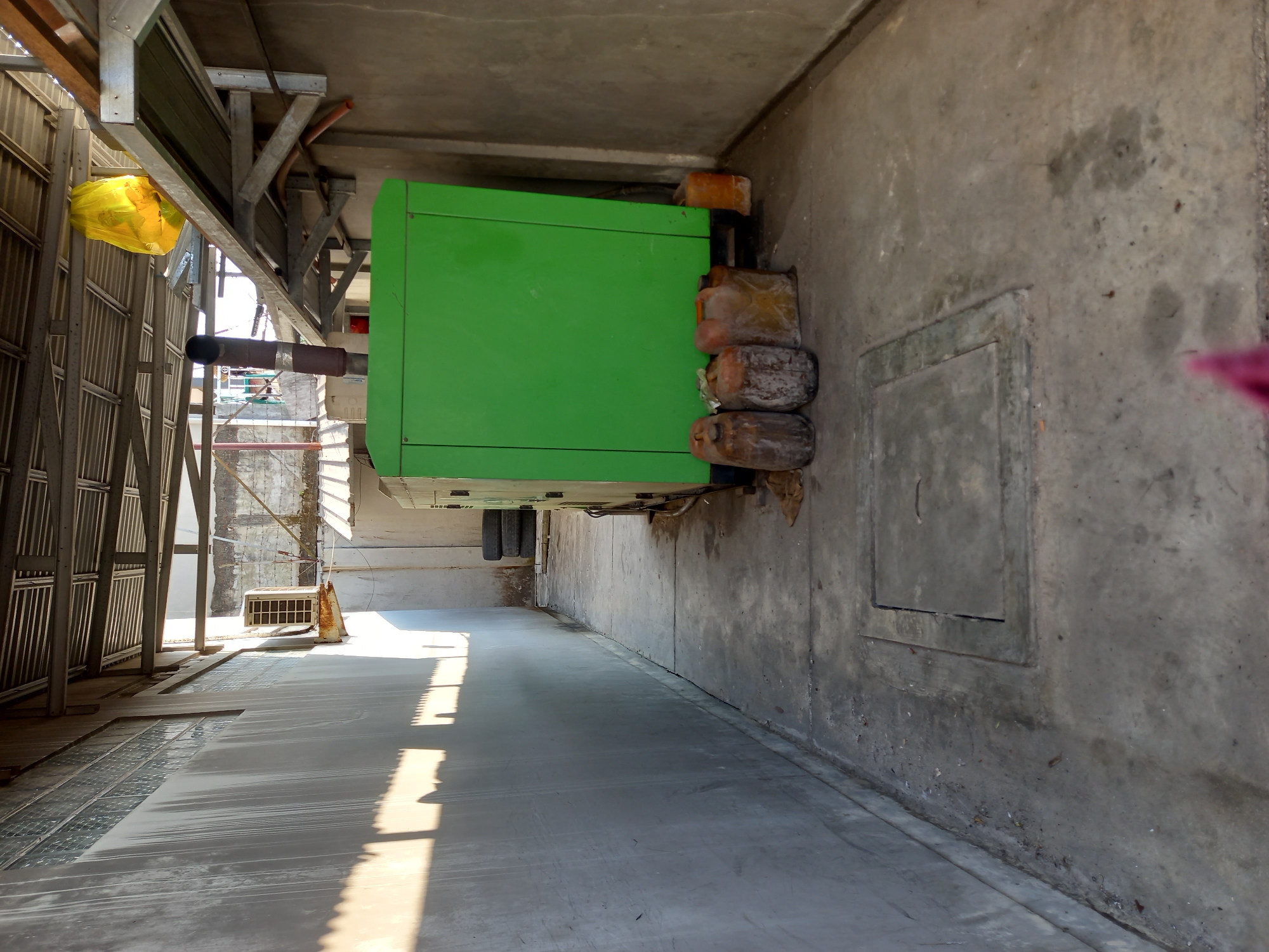 The image depicts an outdoor alleyway in Samarinda, Indonesia, likely a service area behind a building. The alleyway is paved with concrete and has grey concrete walls on both sides. A corrugated metal roof structure extends over the alleyway.
Dominating the right side of the image is a large, bright green generator unit. To the left of the generator, there are several old, discolored plastic barrels, some appearing weathered and dirty, and a small orange plastic container. Behind the generator, a stack of black tires is visible. A metal exhaust pipe extends vertically from the generator.
On the left side of the alley, a long, grey concrete wall features an air conditioning unit mounted high up, showing signs of rust. The wall is illuminated by harsh sunlight, casting strong, elongated shadows across its surface. Further down the alley, a concrete manhole cover is set into the ground.
In the background, beyond the generator, structures with visible windows and what appears to be laundry hanging on a line can be seen. A bright yellow plastic bag is suspended from the metal roof structure on the right. The overall impression is one of a functional, utilitarian space that is somewhat neglected.
