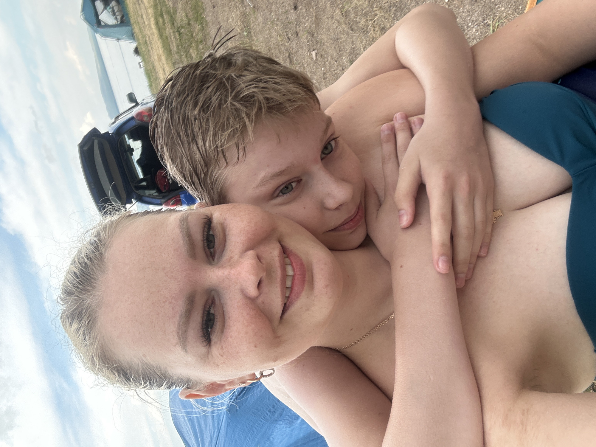 A selfie taken from a low angle shows two individuals, a woman and a younger male, embracing on a beach. The woman, in the foreground, has blonde hair pulled back and is smiling. She is wearing a teal bikini top and a necklace. The boy, positioned behind and to the right of the woman, has brown hair and is resting his head on her shoulder, with his arms wrapped around her. He is looking towards the camera with green eyes.
In the background, a blue car with its trunk open is parked on the grassy shore. Further in the distance, there are tents and a body of water that appears to be a lake or the sea. The sky is overcast with clouds. The location is identified as the city of Жемчужный, Russia, suggesting a lakeside or coastal setting. The overall scene depicts a casual, outdoor moment of affection and leisure.