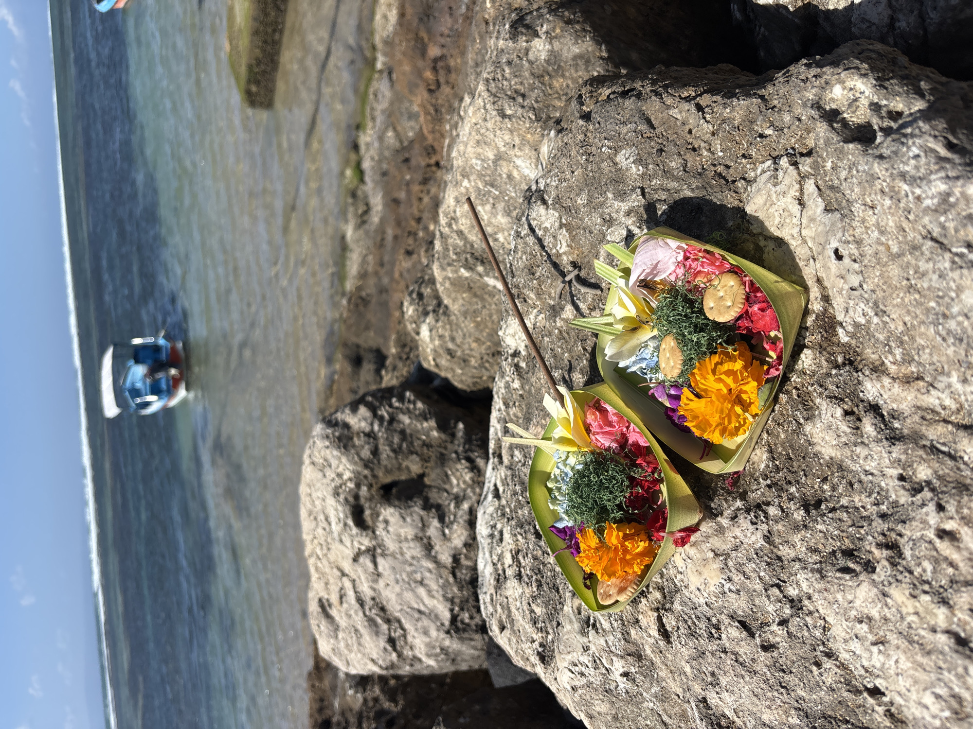 Two small, leaf-lined offering baskets filled with colorful flowers, green foliage, and small cookies are placed on large, rough-textured rocks at the edge of the ocean. A thin, dark incense stick rests across one of the baskets. In the background, the ocean is visible with gentle waves breaking and a small blue and red boat with a white canopy anchored offshore under a clear blue sky. The scene appears to be on a beach or rocky coastline in Denpasar, Indonesia, likely a location used for making offerings. There are no people visible in the image.