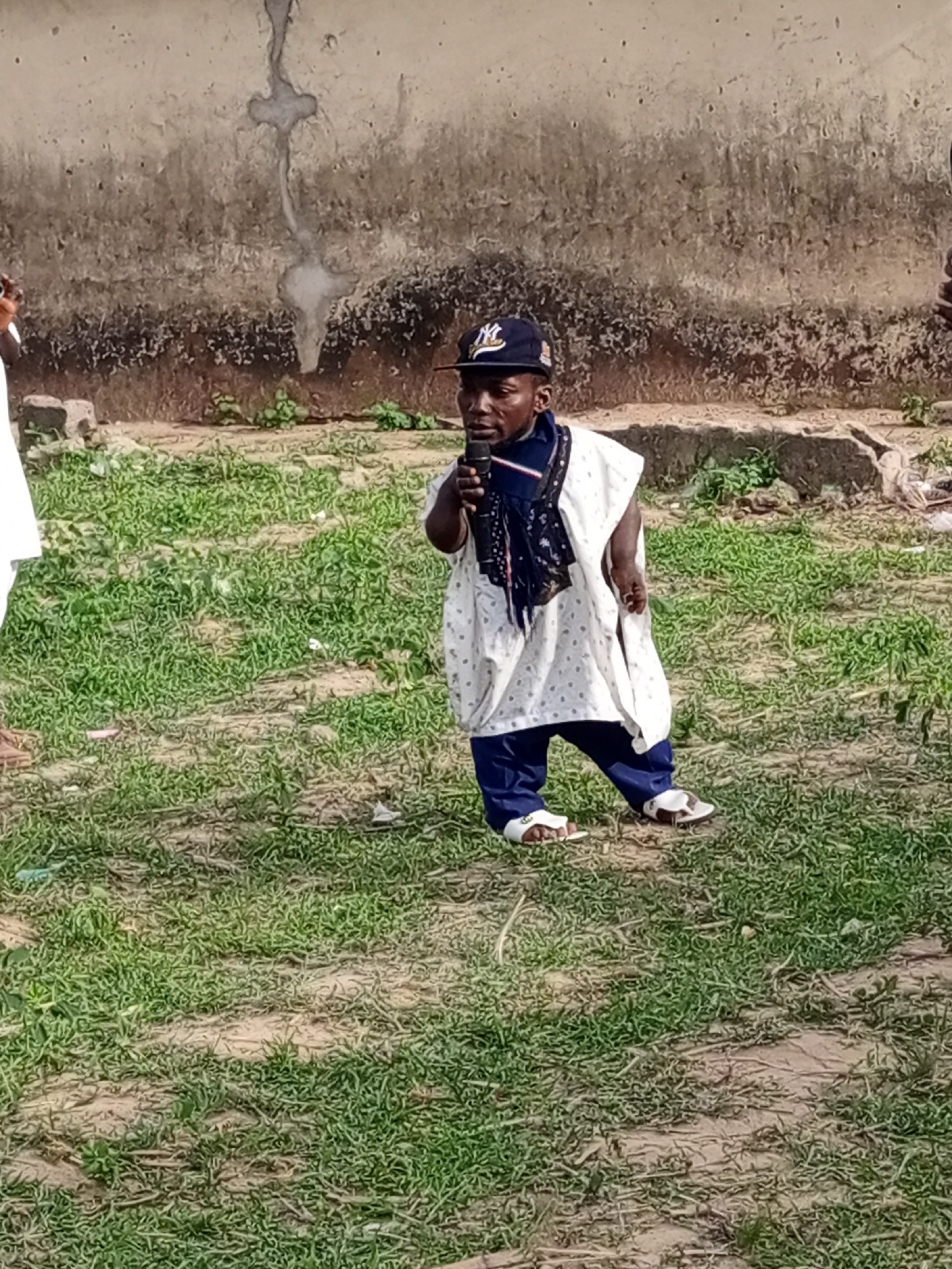 A short-statured man stands on a patch of grass in what appears to be an outdoor setting in Keffi, Nigeria. He is wearing a dark blue New York Yankees baseball cap, a white tunic with patterned blue dots, a dark blue scarf around his neck, dark blue pants, and white sandals. He is holding a microphone in his right hand, positioned close to his mouth. The background shows a weathered wall with stains or discoloration, suggesting an urban or semi-urban environment. The ground is covered with grass, and there are patches of bare earth visible. Parts of other individuals can be seen in the edges of the frame.
