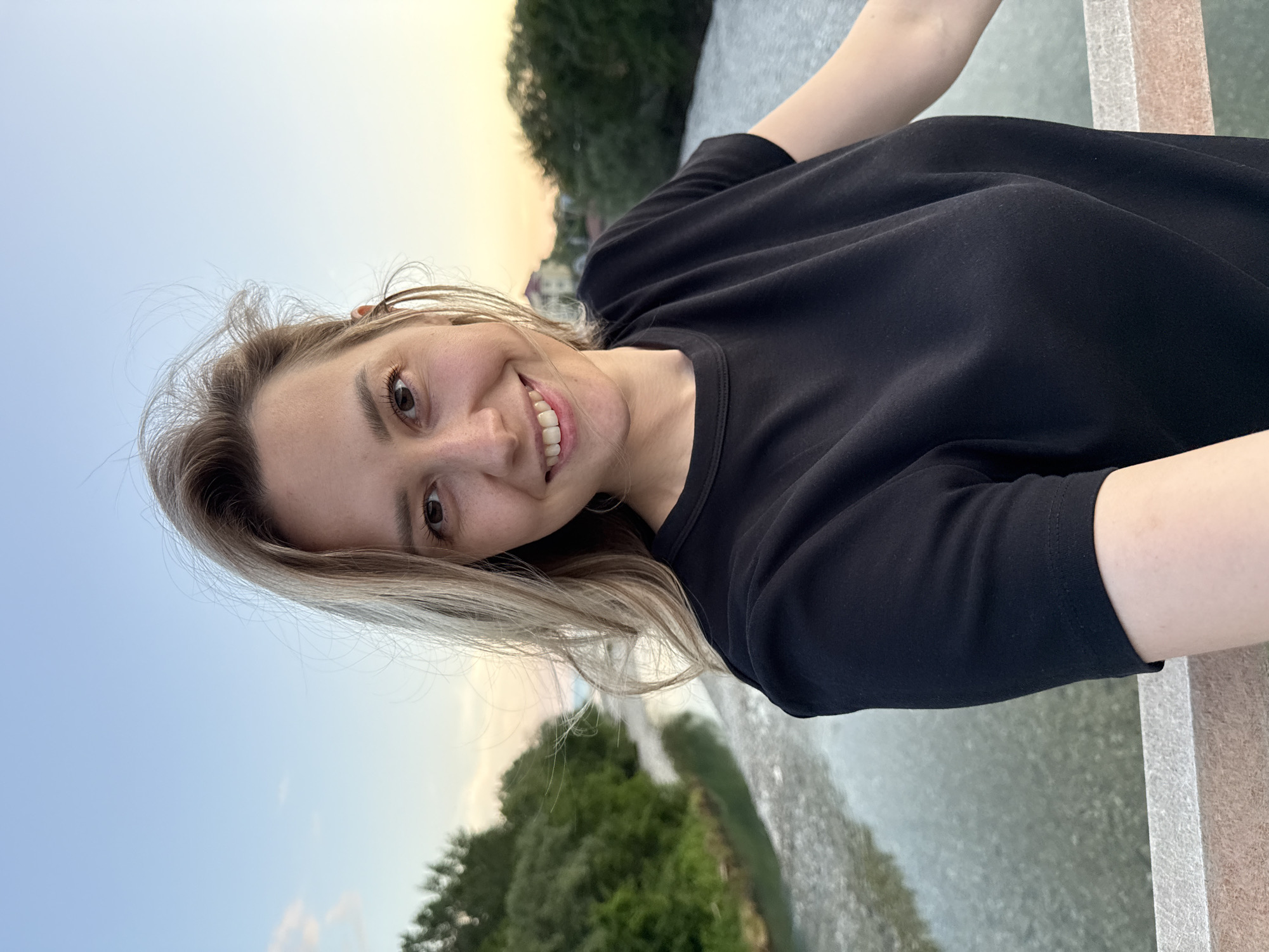 A woman with blonde hair and fair skin is taking a selfie on a bridge. She is wearing a black t-shirt and smiling. In the background, there is a river with trees on either side, and the sky is a pale blue with some clouds. The location is identified as Nebug, Russia.
