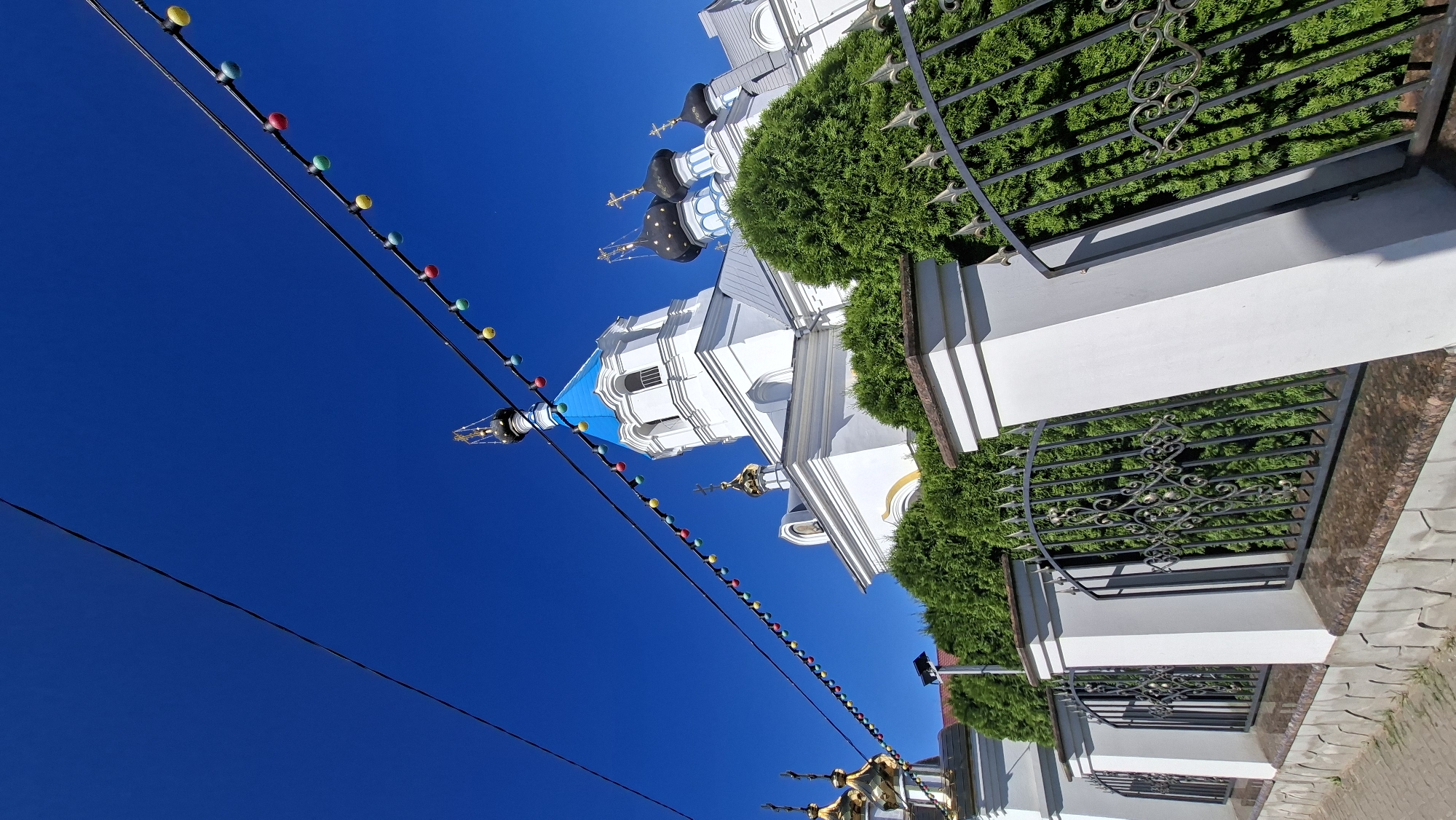 The image shows a view of the Holy Trinity Church in Pastavy, Belarus, on a sunny day. The church has white walls and a blue dome, with smaller golden domes visible on the left. A black metal fence with decorative elements runs along the foreground, separating the viewer from a row of green evergreen bushes. Above the church and fence, a bright blue sky is visible, with power lines strung with colorful lights running diagonally across the frame. The overall scene suggests a festive atmosphere, possibly during a holiday or event.
