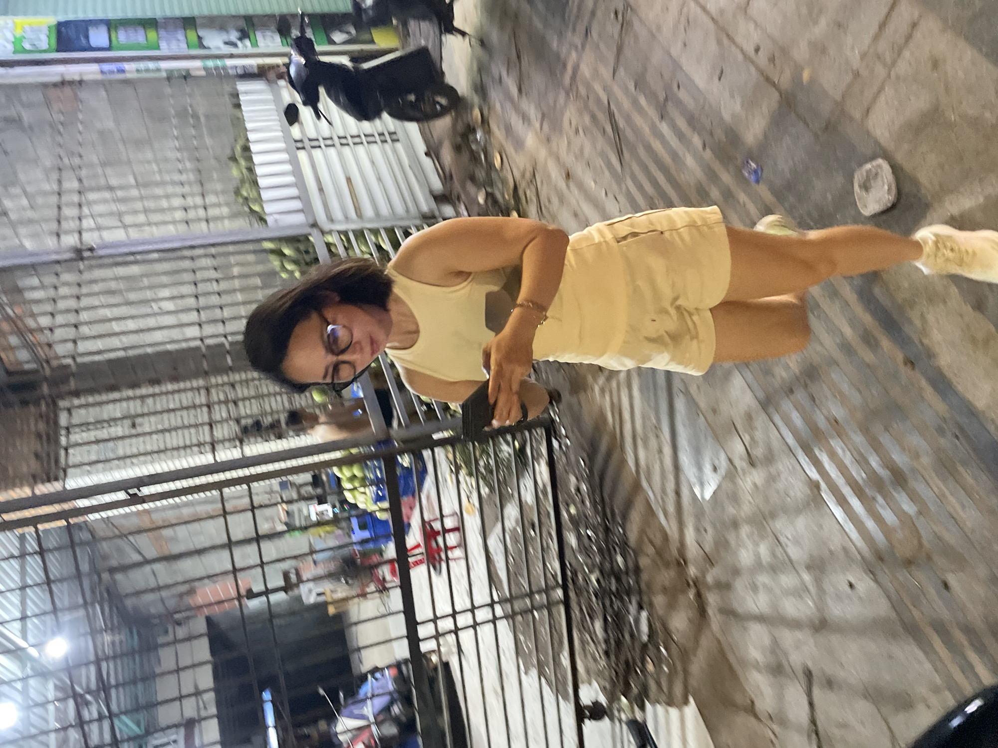 A woman is walking down a sidewalk in Phu Thuy Ward, Vietnam. She is wearing a yellow tank top and yellow shorts, with white sneakers on her feet. She is holding a black cellphone in her right hand and looking down at it. The woman is in her 30s and has short black hair and is wearing glasses. The sidewalk is made of concrete and there is a metal fence behind her. In the background, there are some motorcycles and a building. There are also some green coconuts in a pile on the sidewalk. The scene appears to be a market or a busy street.