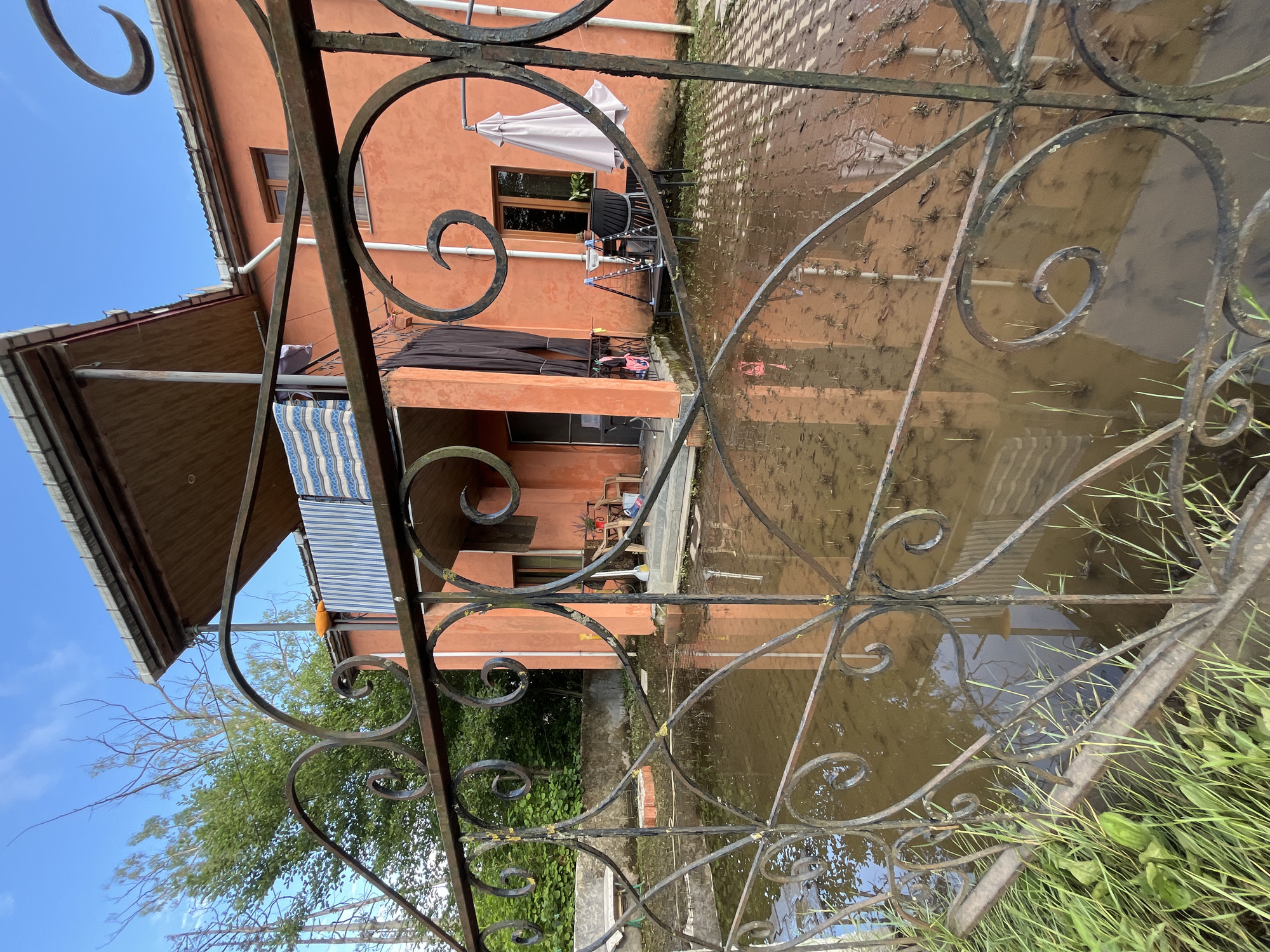The image depicts a flooded courtyard in front of a two-story house with an orange stucco exterior, located in Ureki, Georgia. The foreground is dominated by a dark, ornate metal fence with decorative scrollwork. Beyond the fence, a large area is submerged in murky brown water, reflecting the house and the sky. Small green plants are visible emerging from the shallow water, and some are also growing in the flooded areas.
The house has a covered balcony on the second floor, with laundry hanging on a line. The ground floor shows a tiled patio area partially submerged by the floodwaters. Outdoor furniture, including chairs, a table, and a closed umbrella, are visible on the patio, some of which appear to be affected by the water. A broom and shovel lean against the house near the entrance. The overall impression is one of recent flooding, with the water level significantly altering the appearance of the yard and immediate surroundings. The sky is a clear blue with a few wispy white clouds.