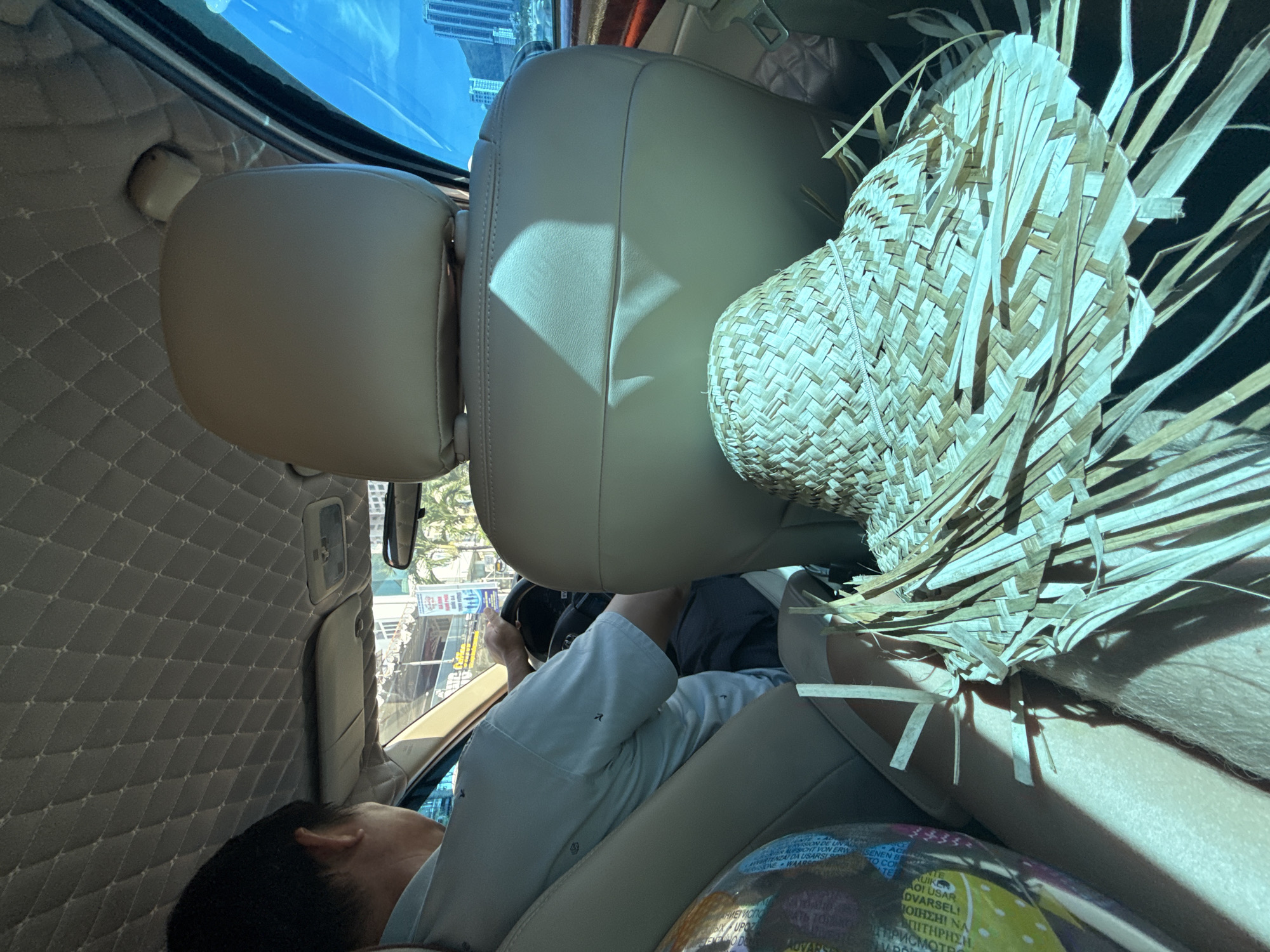 This image shows the interior of a car in Nha Trang, Vietnam, from a rear-facing perspective. A man is driving the car, wearing a light-colored collared shirt and dark pants. His hands are on the steering wheel. In the foreground, on the passenger seat, is a woven straw hat with fringe. A portion of a colorful patterned object, possibly a bag or globe, is visible on the left side of the seat. Sunlight streams into the car, creating shadows on the car's interior, particularly on the back of the driver's seat and the dashboard. The car's interior appears to be upholstered in a light, possibly beige, material. Outside the car windows, buildings and palm trees are visible, indicating an urban environment. The sky is bright blue with some wispy clouds.