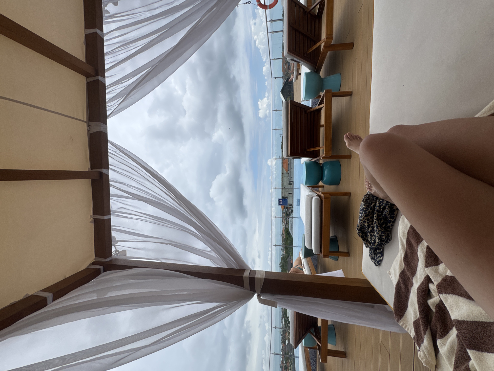 The image depicts a person's legs resting on a lounge chair under a canopy with white sheer curtains. The lounge chair is on a wooden deck next to a swimming pool. In the background, there are more lounge chairs, blue stools, and a railing overlooking a city and the ocean. The sky is cloudy. The scene is located at a rooftop pool area in North Kuta, Indonesia.