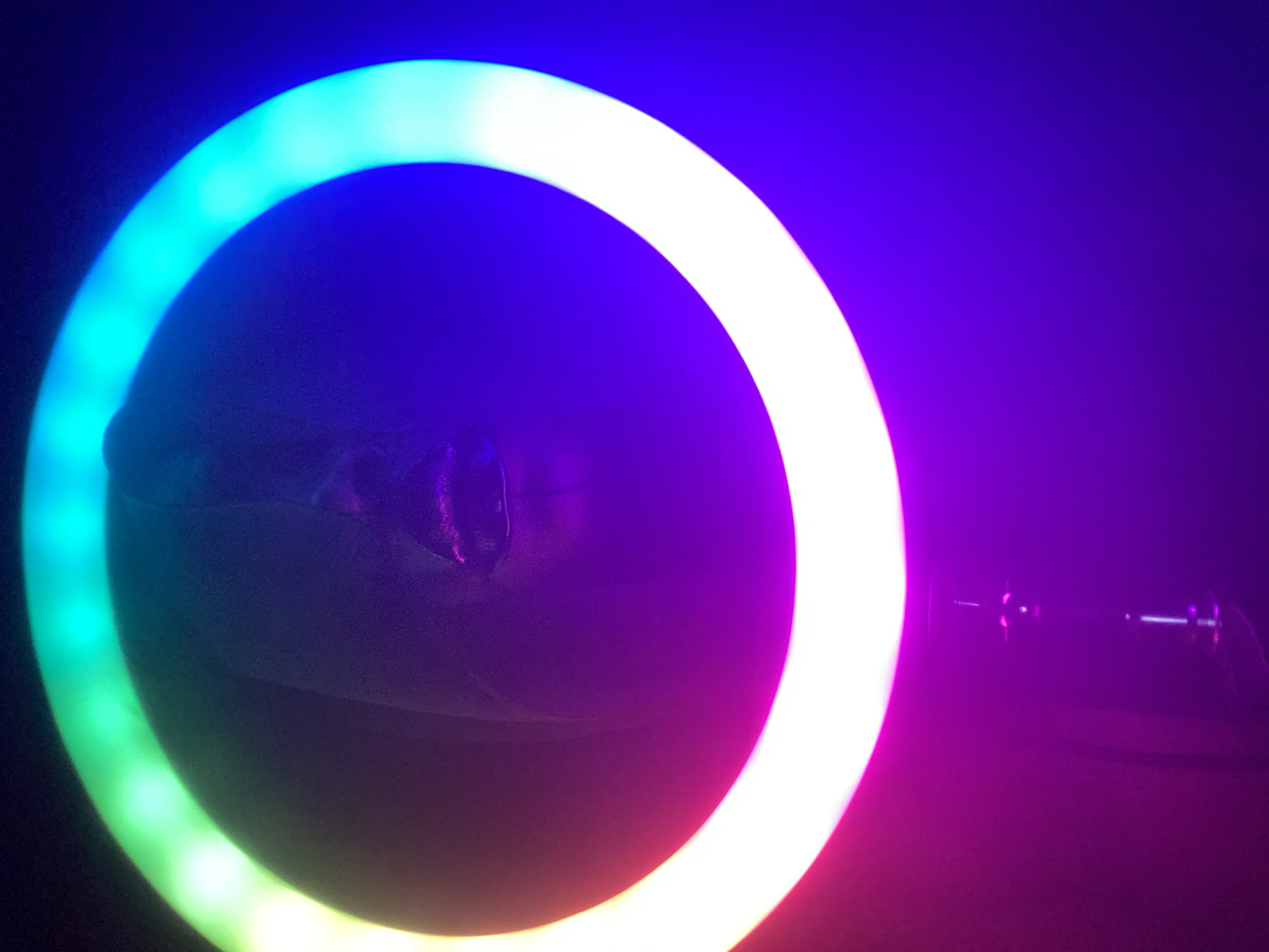 The image shows a person laying down in front of a circular LED light. The light is multicolor, transitioning from green to white to pink. The person is indistinct, appearing shadowed and covered by a blanket or fabric. A portion of the light stand is visible to the right of the ring light. The setting is dark and indistinct, suggesting a dimly lit room. The image was taken in Maigizo, Nigeria.