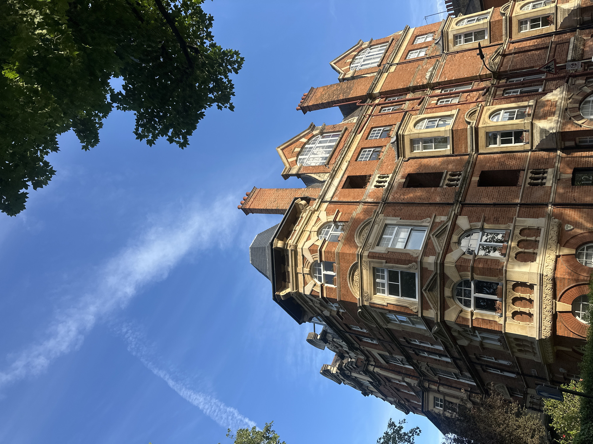 A low-angle view of a red brick building in London, United Kingdom, taken during the day. The building has multiple floors, each with ornate stone detailing around the windows. The architecture is English Baroque. The sky is a clear blue with white contrails. Green leaves are visible in the top left corner and the lower right corner of the image.