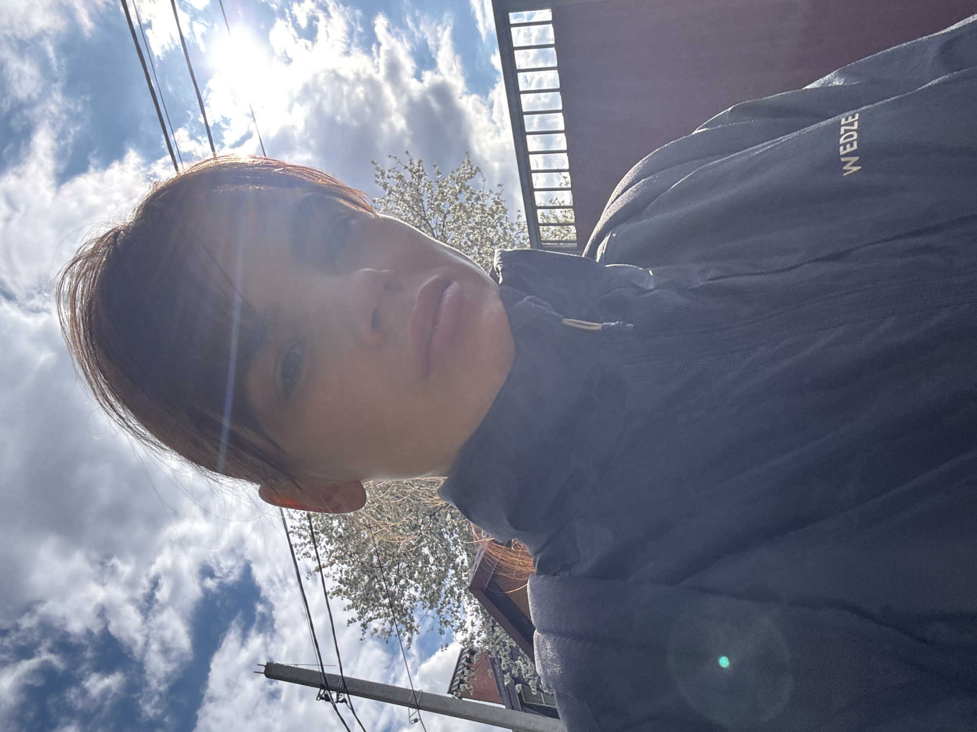 This image is a highly tilted, upward-angled selfie featuring a person outdoors in Rostov-on-Don, Russia. The primary subject is an individual, likely a woman, with dark brown hair and fringe, looking directly towards the camera. Her face is partly in shadow and partly illuminated by strong sunlight, causing lens flare visible near the sun's position. She has a neutral expression with slightly pursed lips.
The person is wearing a dark-colored, possibly padded, jacket or coat. On the right shoulder area of the jacket, the white text "WEDZE" is clearly visible.
The scene is set against a bright blue sky filled with white, fluffy clouds, indicating clear weather. The sun is high, suggesting midday or early afternoon. Several dark power lines traverse the upper part of the frame, leading towards a utility pole in the lower left. In the mid-ground, behind the person, a tree with abundant white blossoms signifies that the photograph was taken during springtime. To the right, a dark, slatted fence or building structure is also partially visible. The overall perspective is dynamic and casual due to the unusual angle.