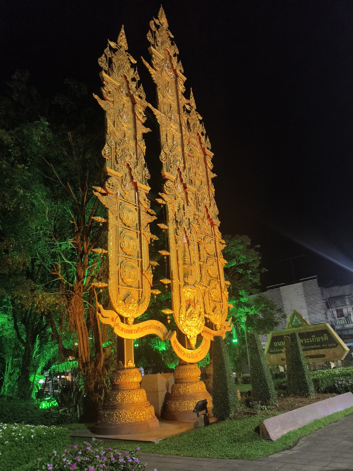 This is a night-time, eye-level, medium shot of an ornate golden monument in Chiang Rai City Municipality, Thailand. The monument, which appears to be two large, towering structures, is intricately decorated with what seems to be religious or cultural motifs. Small, bell-shaped ornaments hang from the main structures. The monument is illuminated, highlighting its golden color and detailed carvings. To the left, trees are brightly lit with green lights, creating a stark contrast against the dark sky. In the background, there are glimpses of buildings, one of which has a distinctive striped facade. A street sign with Thai lettering is visible on the right, indicating the location. The foreground features manicured green grass, hedges, and some small purple flowers. The overall scene suggests a public space or a significant landmark in the city. There are no people visible in the image.