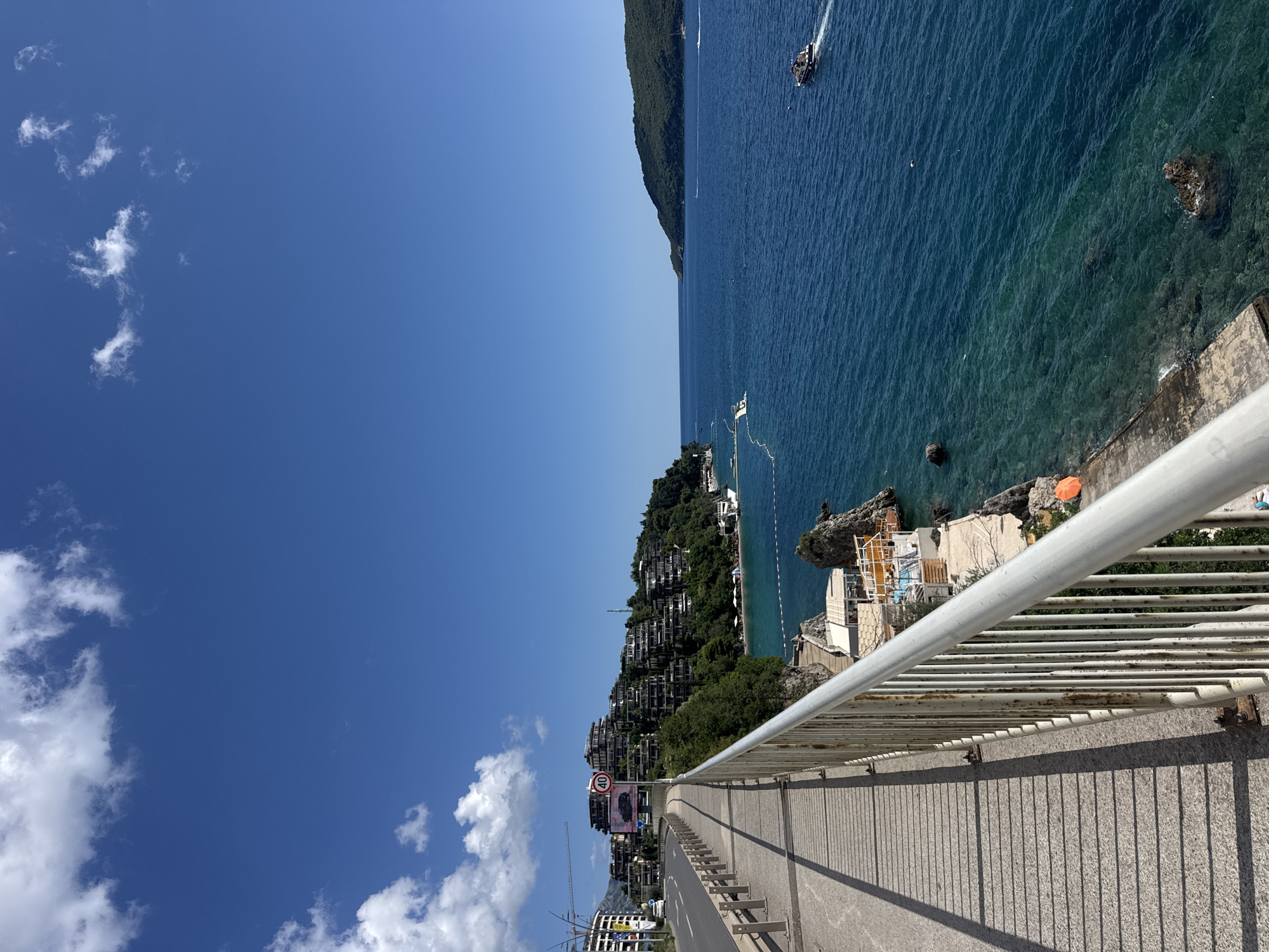 This is a medium-angle, eye-level, vertical shot of a coastal scene in Budva, Montenegro, under a clear blue sky with scattered clouds. In the foreground, a white railing runs along the edge of a concrete walkway, casting long shadows from the midday sun onto the textured pavement. To the left of the walkway, a road curves uphill, with a white car visible and a speed limit sign indicating 40. Buildings with rounded, stepped architecture are built into the lush green hillside overlooking the water. The clear, turquoise sea is in the middle ground and background, with a rocky coastline visible on the right, featuring a small beach area with lounge chairs and an orange umbrella. Several boats are present in the water, including a white speed boat and a larger passenger boat moving away from the shore. A distant, tree-covered hill forms the horizon. The scene suggests a warm, sunny day, likely in a resort area.