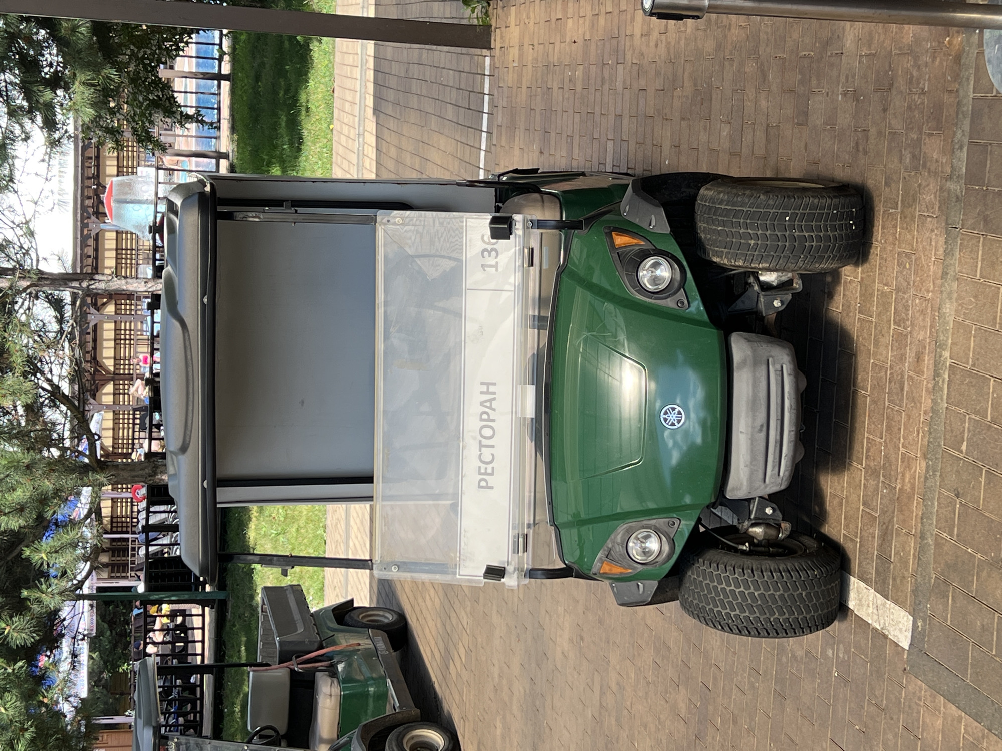 This image shows a green Yamaha golf cart parked on a brick-paved walkway. The golf cart has a clear windshield with a sign that reads "РЕСТОРАН" (Restaurant) in Russian and the number "136". There is another golf cart visible in the background to the left. Behind the golf carts, there are trees, a fence, and what appears to be a swimming pool area with people in the distance. The environment suggests an outdoor recreational or resort location. The specific location in the city of Plast, Univska, Russia, is not explicitly discernible from the image itself, but the presence of a golf cart and pool area suggests a leisure facility.