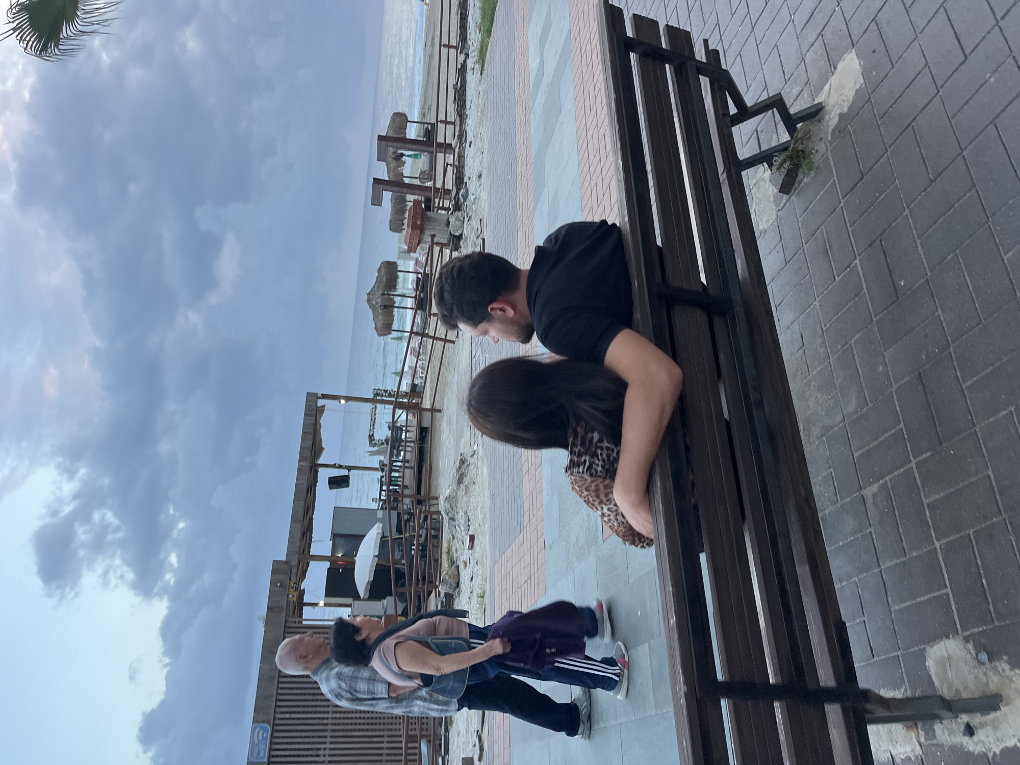 The image shows a man and a woman sitting on a wooden bench by the sea in Alanya, Turkey. The man is wearing a black shirt. The woman has long, dark hair and is wearing a leopard print top. They are seated next to each other. Behind them are beach structures with thatched roofs. Further in the background is the sea and a cloudy sky. To the left of the couple are three other people standing in front of a building with a blue sign. The foreground is a brick and tile pathway where the bench sits.