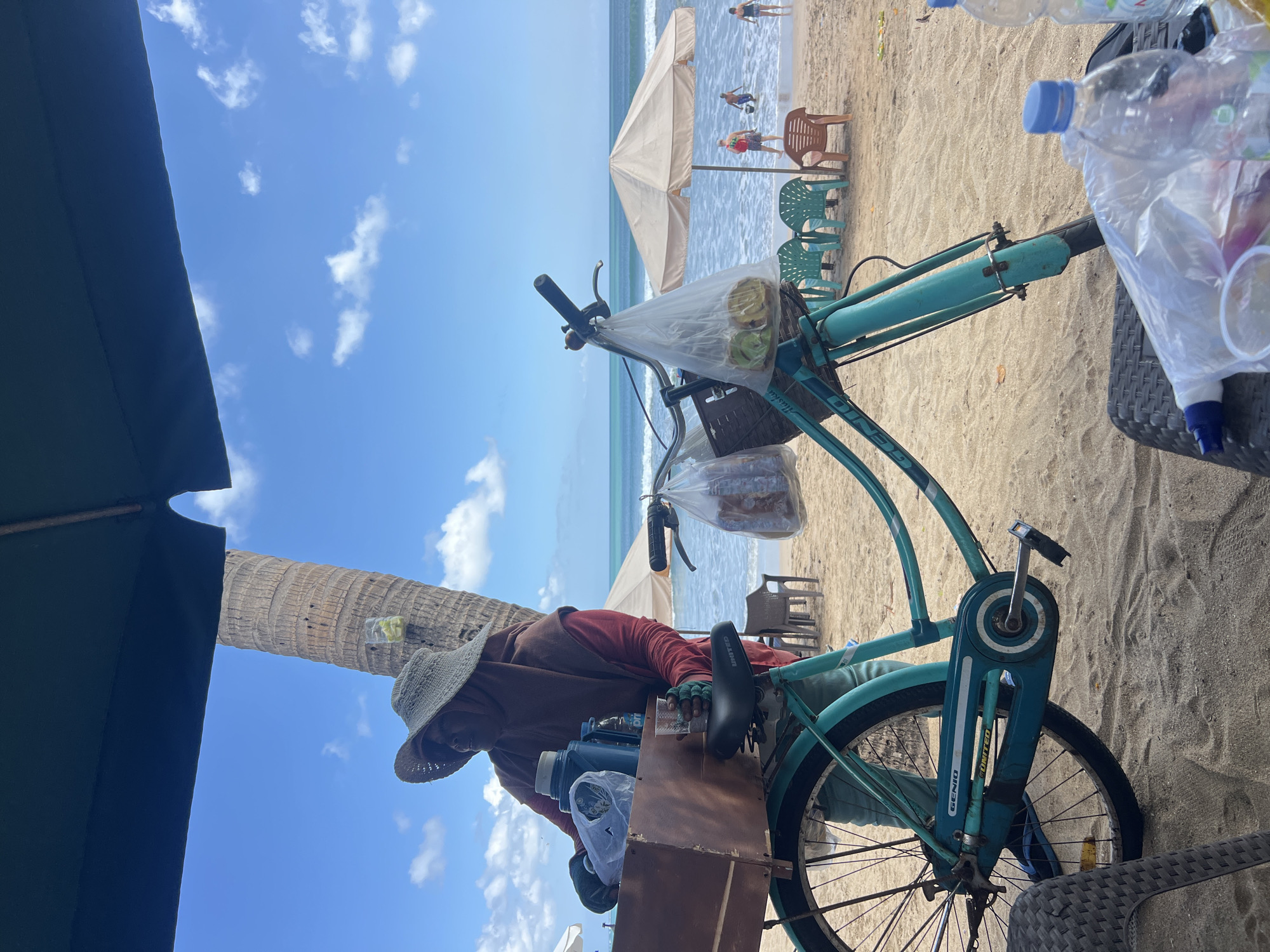 The image depicts a beachfront scene in Kuta, Indonesia. A person wearing a hat and a headscarf stands next to a teal bicycle. The bicycle has a wooden crate attached to the rear, containing a blue thermos and plastic bags of goods. There are also plastic bags hanging from the handlebars, one containing what appears to be food items. In the background, beach umbrellas and chairs are set up on the sand, with the ocean and a blue sky with white clouds visible. Several people are seen in the water. The overall atmosphere suggests a vendor preparing to sell items on a sunny day.