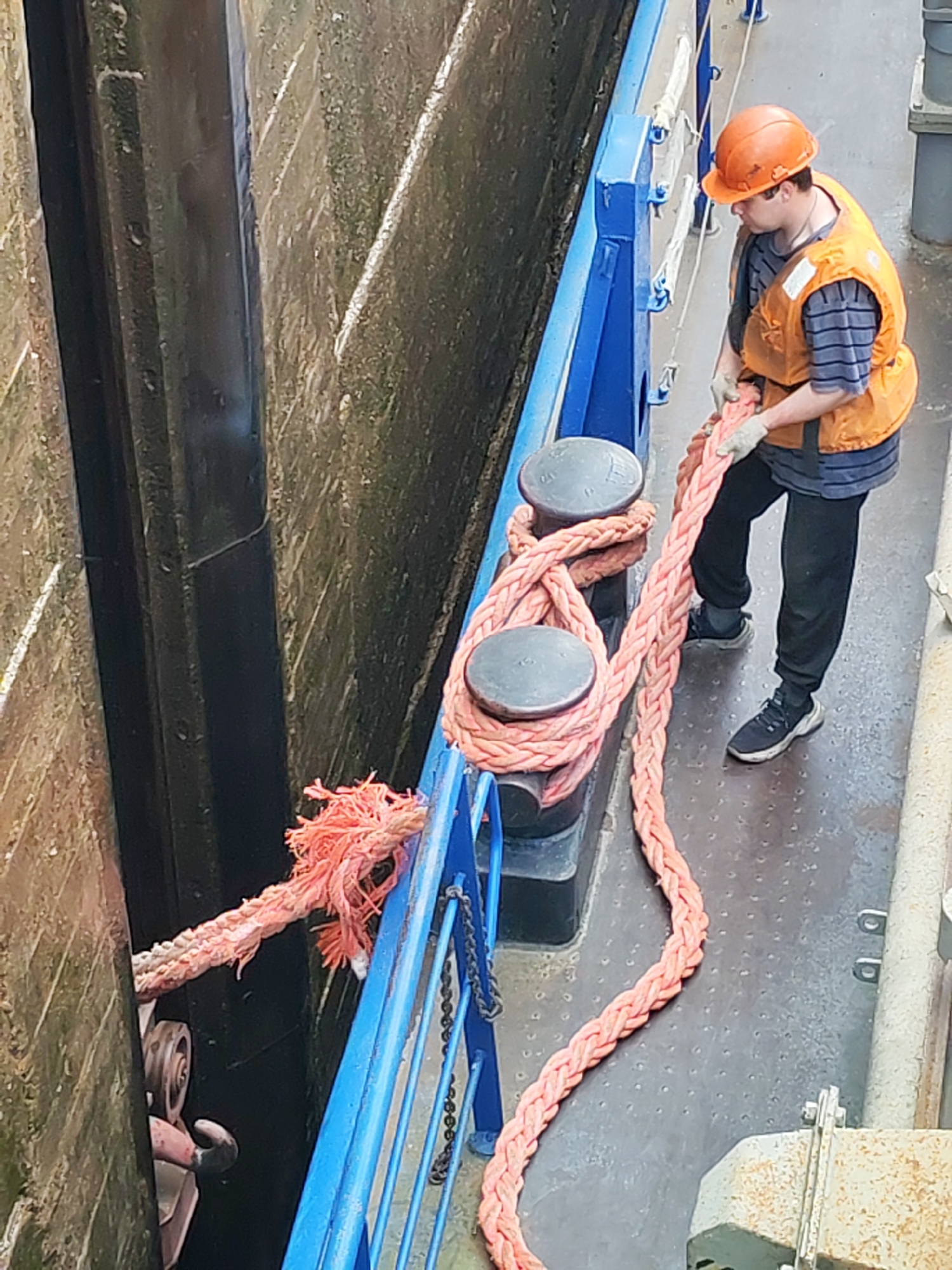 The image depicts a man on a boat in a lock on the Volga-Baltic Waterway in Devyatiny, Russia. The man, wearing a hard hat, safety vest, and gloves, is holding a thick pink rope that is secured to bollards on the boat. The rope extends to the lock wall. The lock walls are made of concrete. A blue railing runs along the side of the boat.