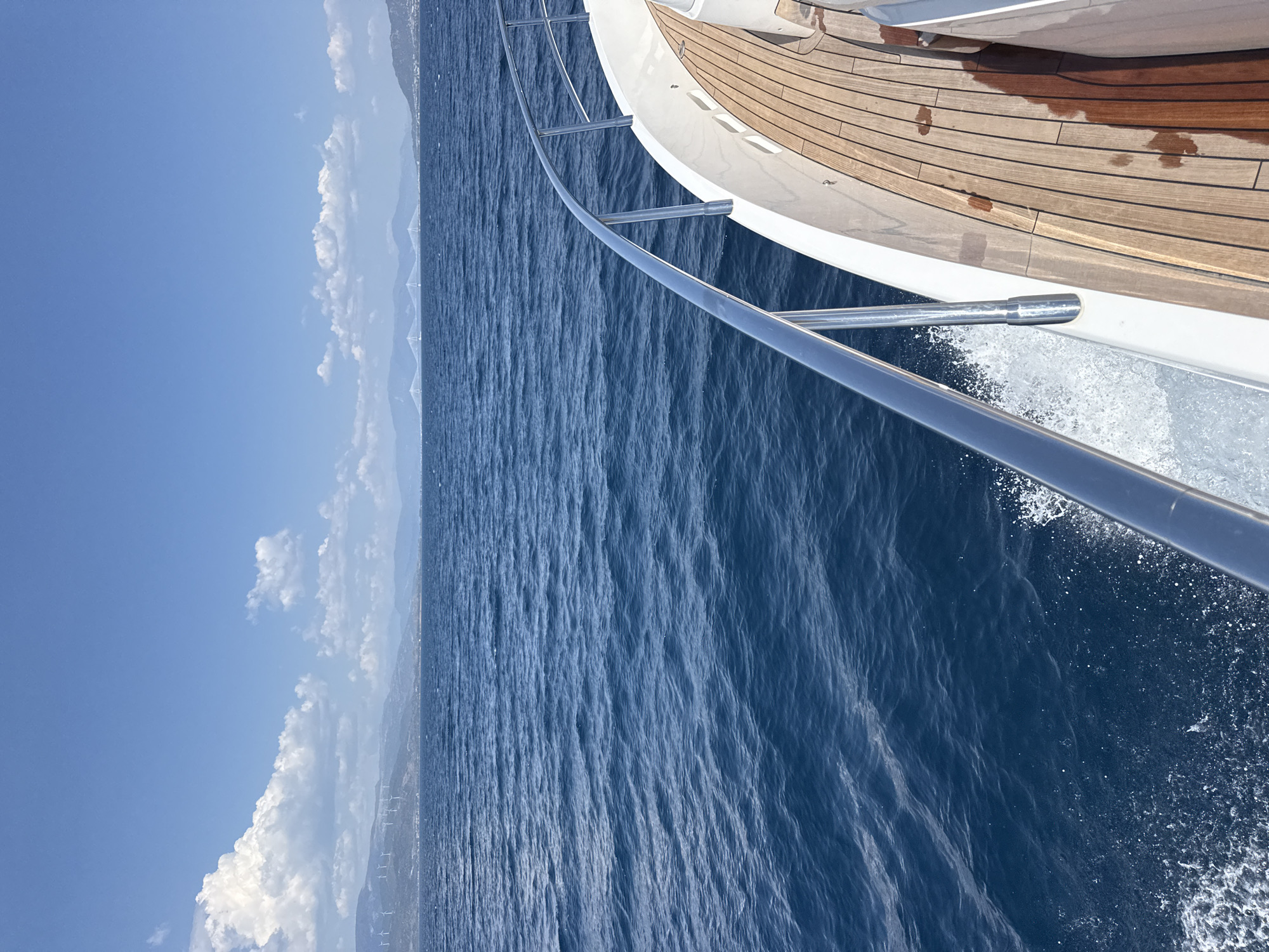 This media file captures a view from the deck of a boat moving through the ocean. The camera is angled downwards towards the water, with the railing of the boat prominently in the foreground. The water is a deep blue with visible ripples and some white spray along the side of the boat, indicating movement. The deck of the boat is made of polished wooden planks, some of which appear wet.
In the distance, a hazy coastline is visible, featuring hills or mountains. There are also several white, vertical structures that resemble wind turbines on the distant land. The sky is a clear, bright blue with scattered white clouds, suggesting a sunny day.
The overall impression is one of a serene journey at sea, likely during daylight hours, given the bright sunlight and clear blue sky. The scene is located off the coast of Patras, Greece, as indicated by the specific details of the landscape and potential wind farms. No people are visible in the frame, and there is no visible text.