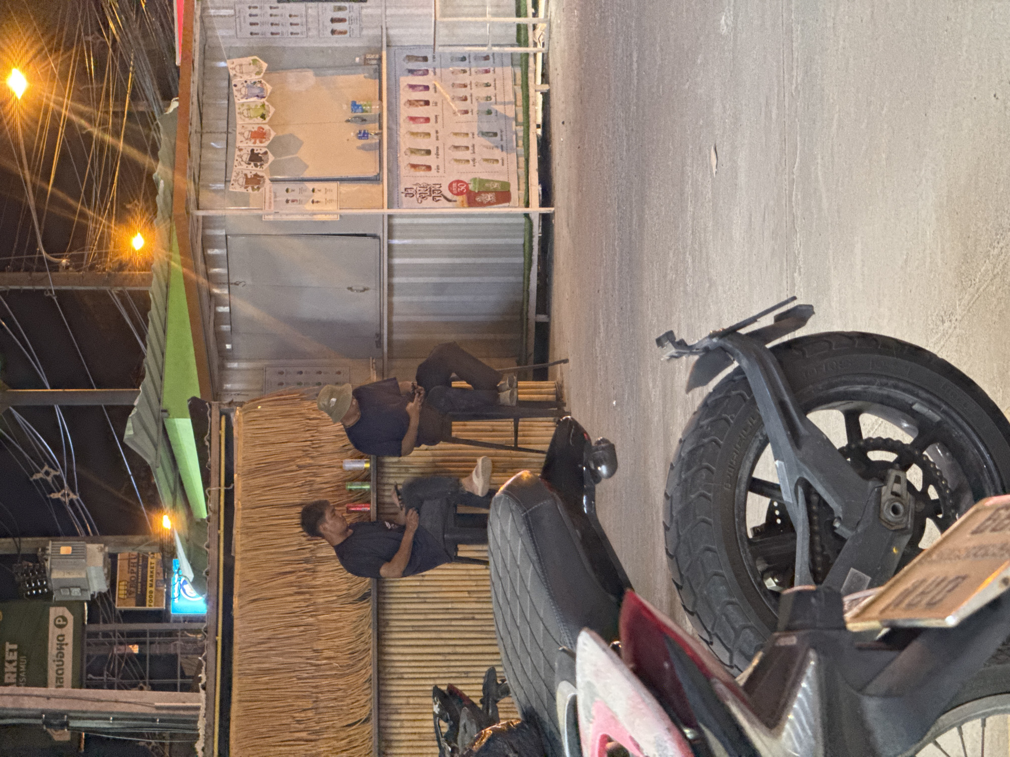 This is an outdoor medium shot, taken at night, of a street scene. The dominant objects are a motorcycle in the foreground and a small stall or shop in the background. Two men are visible, seated on stools in front of the stall. One man, wearing a hat and a dark t-shirt, is looking down at his phone. The other man, in a dark t-shirt and jeans, is also looking down, possibly at his phone or something on the ground.
The stall appears to be a food or drink vendor, with colorful posters displayed on its exterior. One poster shows pictures of various drinks, and another displays a price of "30". The stall has a thatched-like wall and a corrugated metal roof. The ground is paved with concrete. Overhead, numerous wires and lights are visible, suggesting an urban or semi-urban environment. The lighting is artificial and casts a warm glow on the scene, consistent with nighttime. The overall mood appears casual and relaxed, with the two men seemingly taking a break or waiting.
