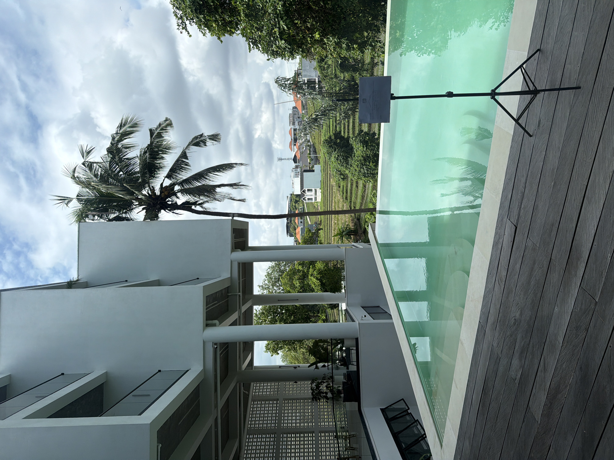 The image depicts a view from a modern building in Mengwi, Indonesia, looking out towards terraced rice paddies and houses. A tall palm tree stands next to the building. A swimming pool with light green water is visible on the right, with a sign on a stand positioned beside it. The pool surface reflects the sky, palm tree, and surrounding landscape. The foreground features dark wooden decking next to the pool and the architectural details of the white building, including columns and a decorative patterned wall. The sky is overcast with heavy cloud cover.