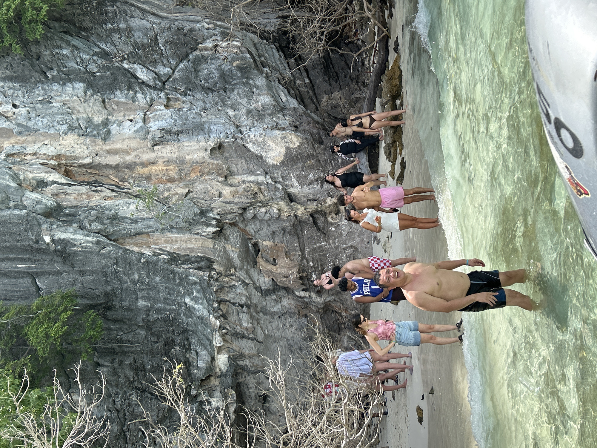 A group of people are gathered on the sandy shore of Haad Hin Khom, Thailand, with a large, weathered cliff face forming the backdrop. The water in the foreground is clear and shallow, with small waves lapping at the shore. The beach is dotted with rocks and driftwood, and sparse, dry vegetation clings to the cliff. Most individuals are dressed in beachwear, with some standing in the water and others on the sand. The mood appears relaxed and casual, with some people interacting and others observing the scenery.