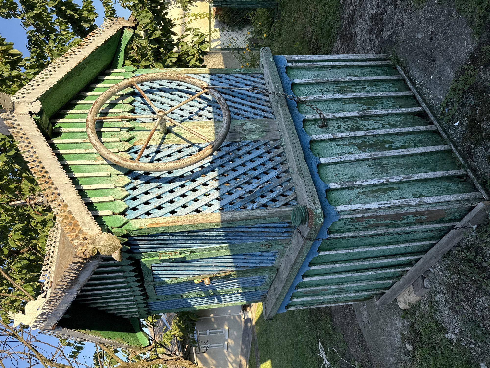 An abandoned, weathered wooden well structure stands in an overgrown yard, bathed in the warm, late afternoon sun. The well's octagonal base is constructed from vertical planks, painted in alternating bands of faded green and white, with a chipped blue trim along the top. Above the base, the structure transitions to a more decorative design with a lattice-like enclosure, painted in blues and greens. A rusty metal wheel, likely part of the well's original mechanism, is mounted on this lattice section. The roof, also in green, features ornate, possibly metal, decorative trim along its edges, including a carved deer figure at the peak. A chain hangs from the wheel, connecting to a coiled rope below. The surrounding area is overgrown with grass and weeds, with a concrete path visible near the well's base. In the background, a modest house with white siding and two windows can be seen, partially obscured by the well and trees. The overall impression is one of neglect and the passage of time in a rural or semi-rural setting.
