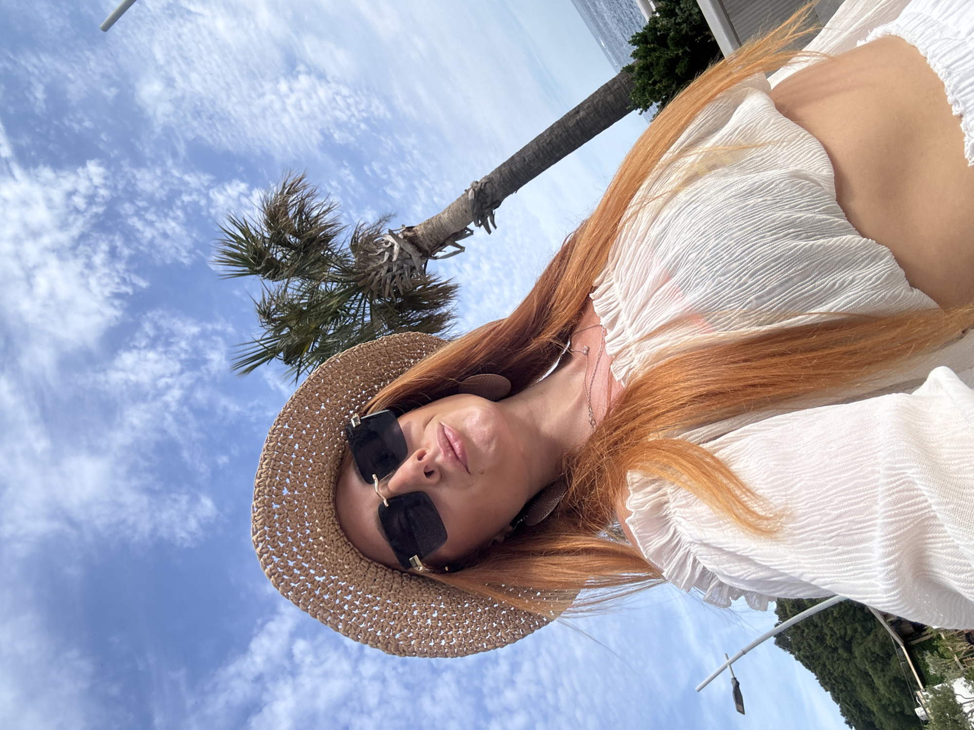 A red-haired woman poses for a selfie, looking slightly upwards towards the camera. She wears a straw hat, dark rectangular sunglasses with gold-toned accents, and large, light-colored, possibly leather, earrings. Her lips are slightly puckered. She is dressed in a white, textured, off-the-shoulder crop top, revealing her midriff, with a small pendant necklace visible. In the background, under a blue sky with scattered white clouds, a tall palm tree stands on the right, overlooking a calm, dark blue sea. A white railing and green foliage are present near the sea. On the left, a light pole is visible against a backdrop of a distant, tree-covered green hill. Indistinct structures are partially visible at the base of the hill. The scene is consistent with a coastal setting in Petrovac centar, Montenegro.