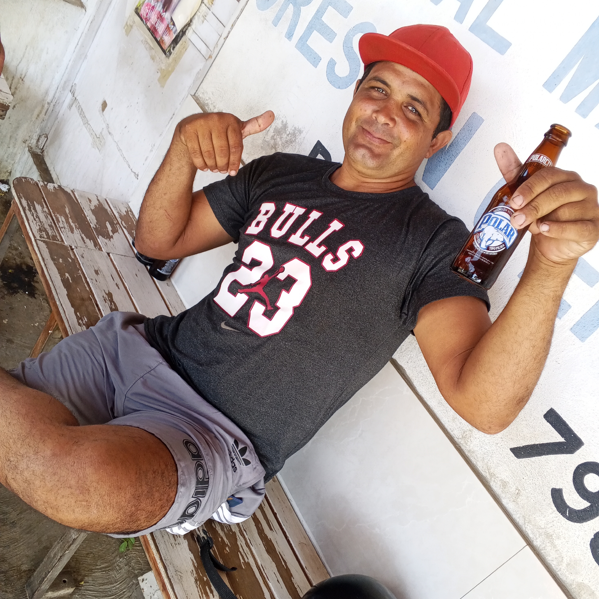 A man wearing a red baseball cap and a gray "Bulls 23" t-shirt sits on a weathered wooden bench. He is also wearing gray Adidas shorts. He holds a bottle of Polar beer in his right hand and points with his left. The background features a white wall with faded blue letters, possibly part of a sign, and large black numerals "79". The location is Unknown, Unknown.