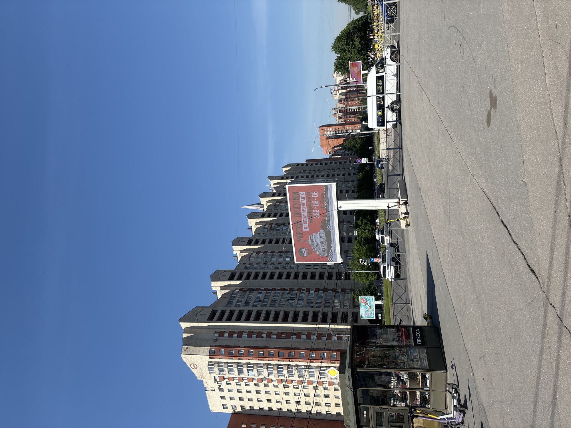 The image captures a bright, sunny urban scene under a clear blue sky. In the foreground, a wide expanse of asphalt pavement shows cracks and faint marks. To the right, a small kiosk with "ПРЕССА" (PRESS) written on its side stands, with several electric scooters parked nearby.
In the midground, a prominent, multi-story building with a distinctive stepped roofline dominates the left side. It features a light-colored facade with sections of red-brown brickwork. Adjacent to this building, a large red billboard displays white Cyrillic text that reads "МОРСКАЯ ПРОГУЛКА ПО ФОРТАМ КРОНШТАДТА" (SEA TRIP TO KRONSTADT FORTS) and a phone number "8 (812) 382-08-08", along with an image of a boat. This indicates the location is likely Saint Petersburg, Russia, given the "812" area code and the reference to Kronstadt.
Beyond the asphalt area, a street is visible with cars, a white van parked next to a metal fence, and traffic lights. A smaller blue billboard and a yellow diamond-shaped road sign (indicating a priority road) are also present. In the background, green trees line the horizon, and a large group of people wearing yellow shirts or jackets can be seen, possibly part of an organized event or tour, near what appears to be a body of water or a river in the far distance. The short shadows suggest it is midday or early afternoon.