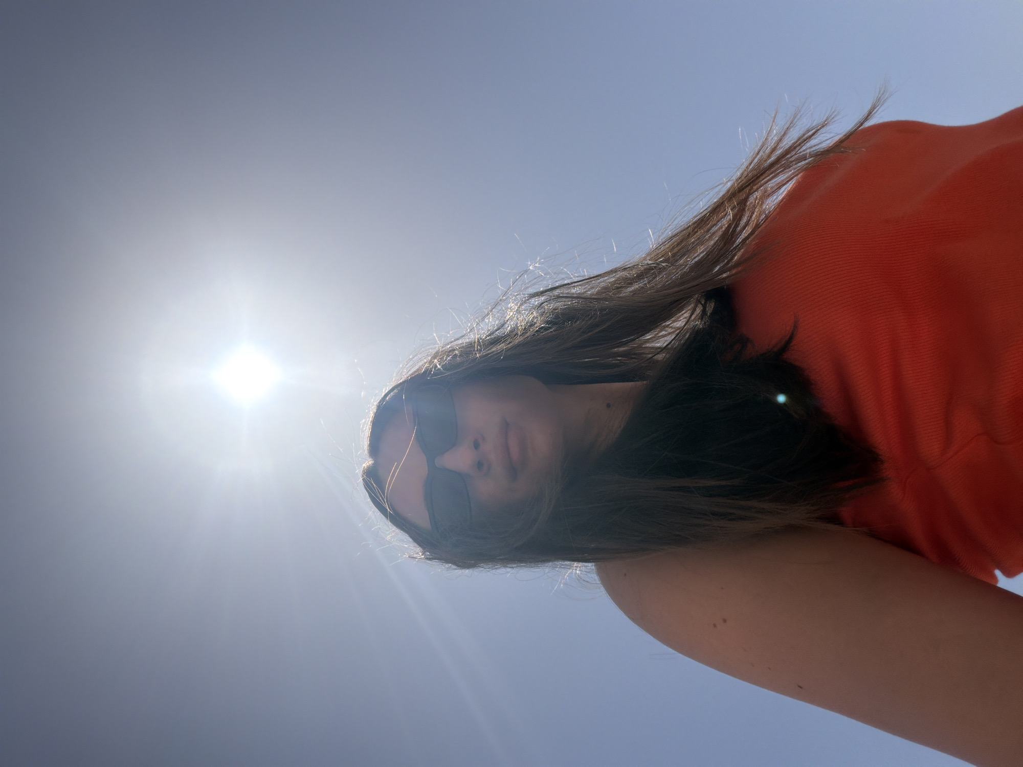 In Sharm El Sheikh, Egypt, a woman is pictured from a low angle outdoors under a bright, clear blue sky. The sun is high and bright, casting lens flare across the image. The woman, with long, dark brown hair that is blowing in the wind, is wearing sunglasses and an orange top. Her left arm and shoulder are visible in the foreground. The background is completely clear sky, indicating an open, outdoor location.